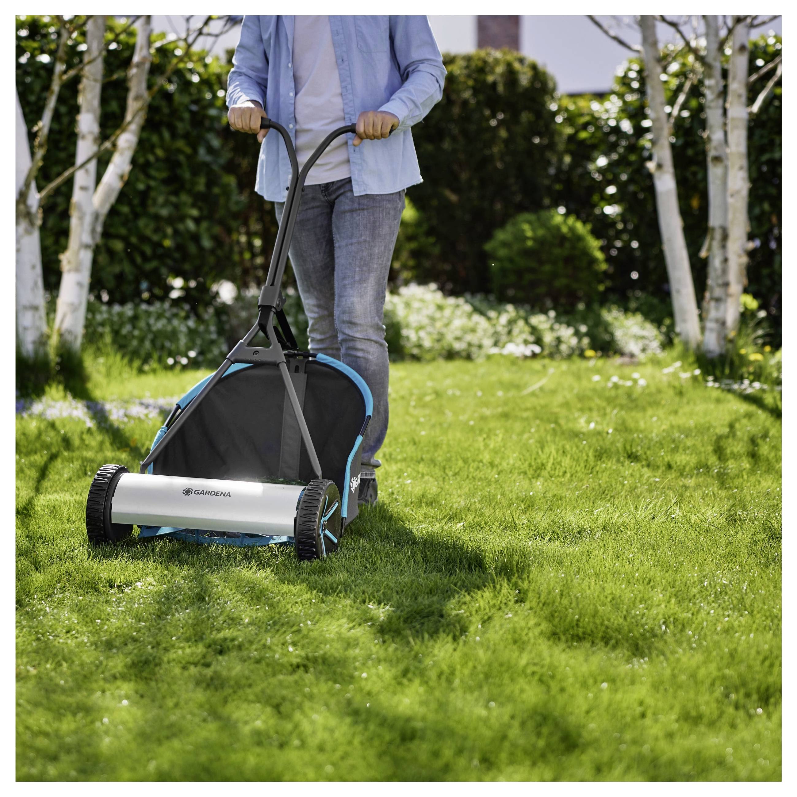 Een persoon gebruikt een handmatige duwgrasmaaier op een zonnige dag. De maaier beweegt zich over een grasrijk gebied, met bomen en struiken op de achtergrond.