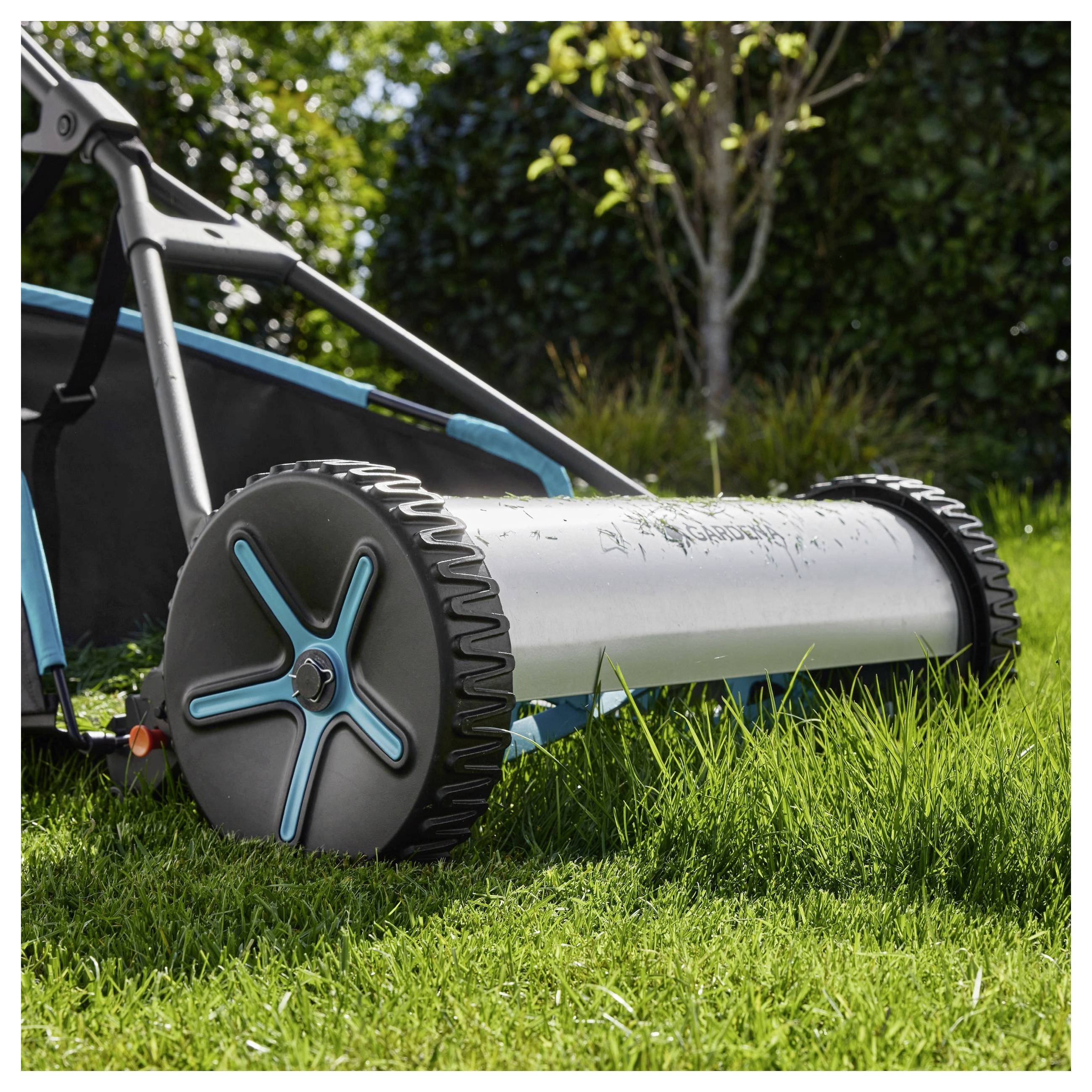 'Een handmatige grasmaaier met blauwe en zilveren details die gras maait op een zonnige dag, met bomen en een haag op de achtergrond.'