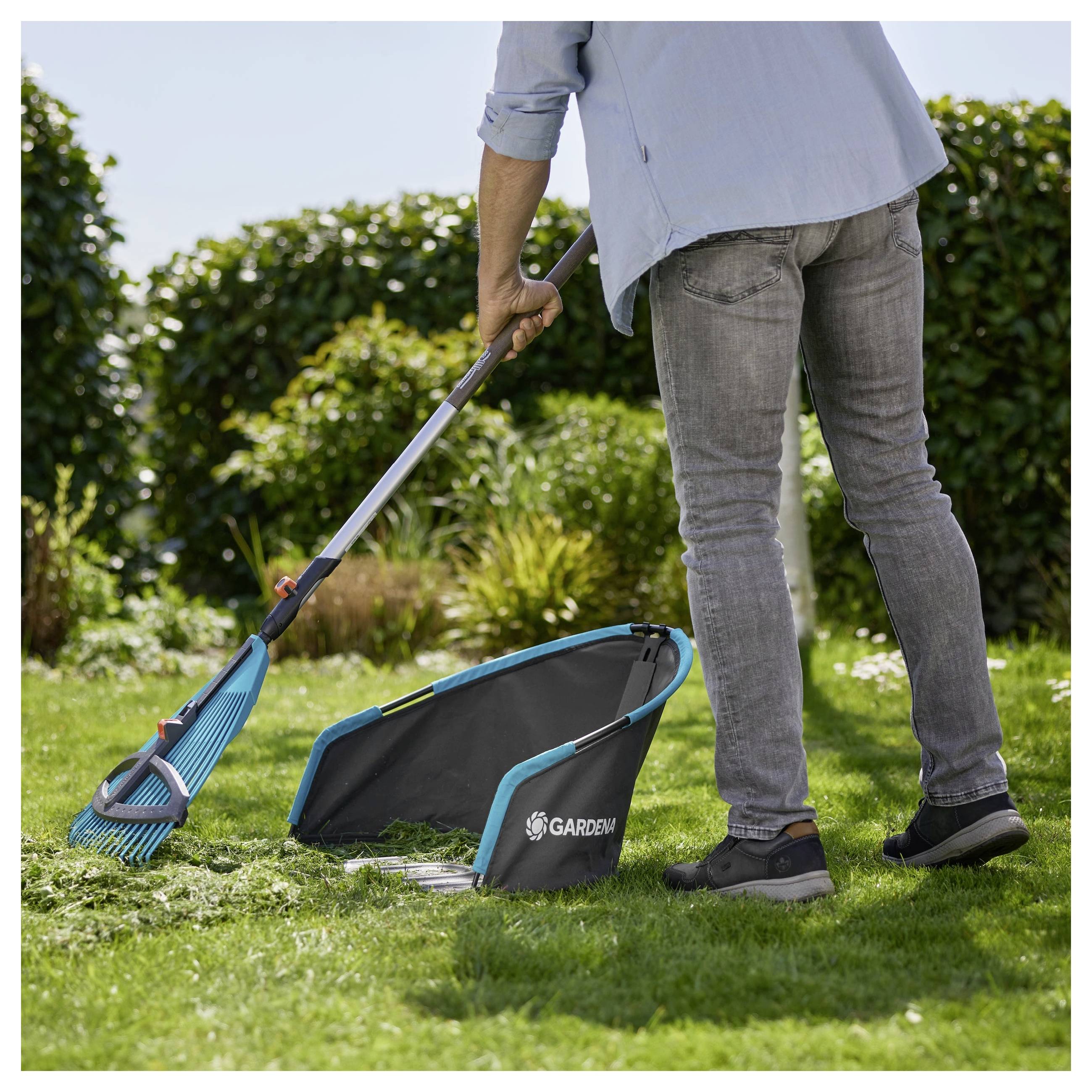 Een persoon harkt gras in een verzamelzak met het label 'Gardena' in een tuinomgeving, omgeven door groene struiken en een heldere blauwe lucht.