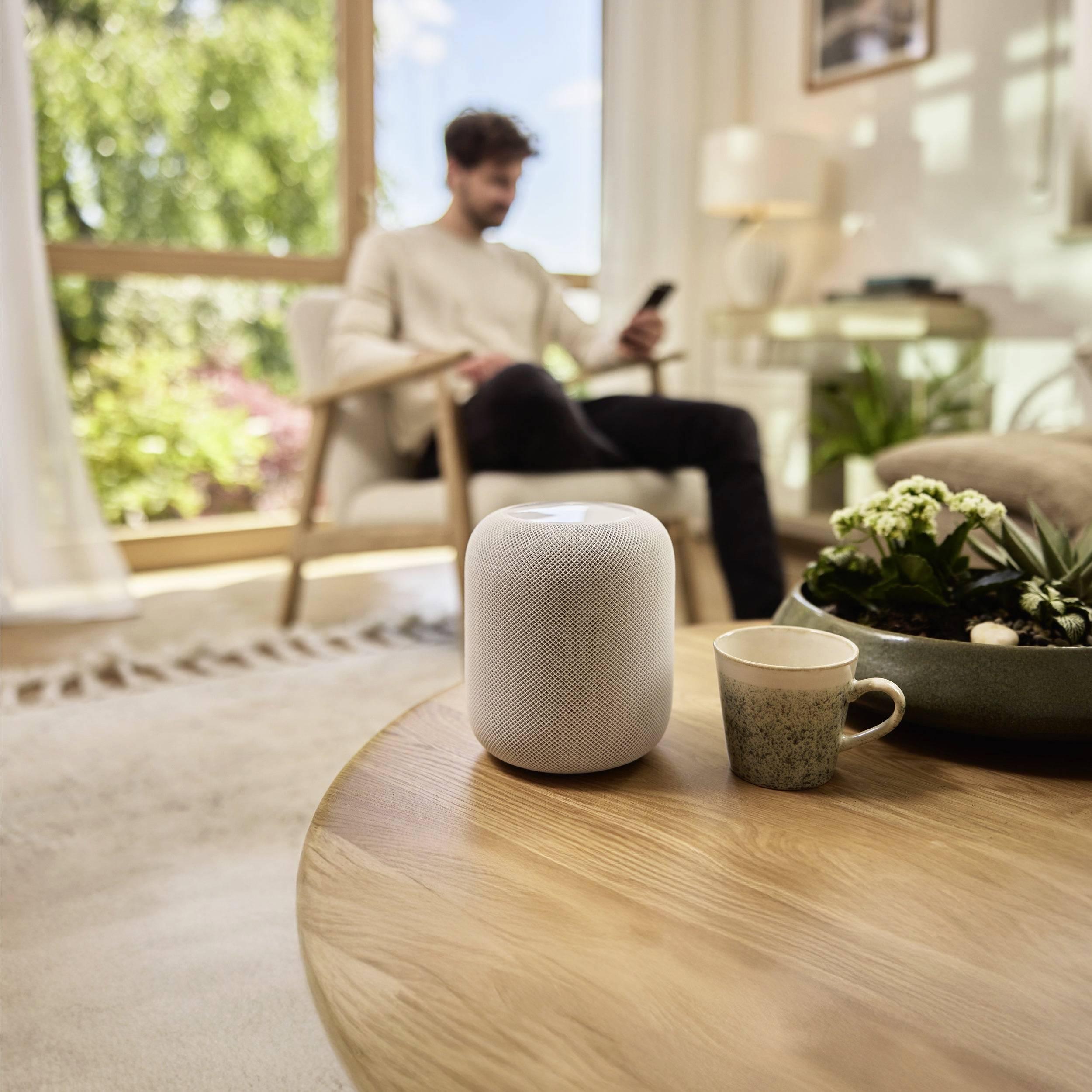 Man in een gezellige woonkamer die een smartphone gebruikt, met een slimme speaker en koffie op tafel; zonlicht stroomt binnen door een raam.