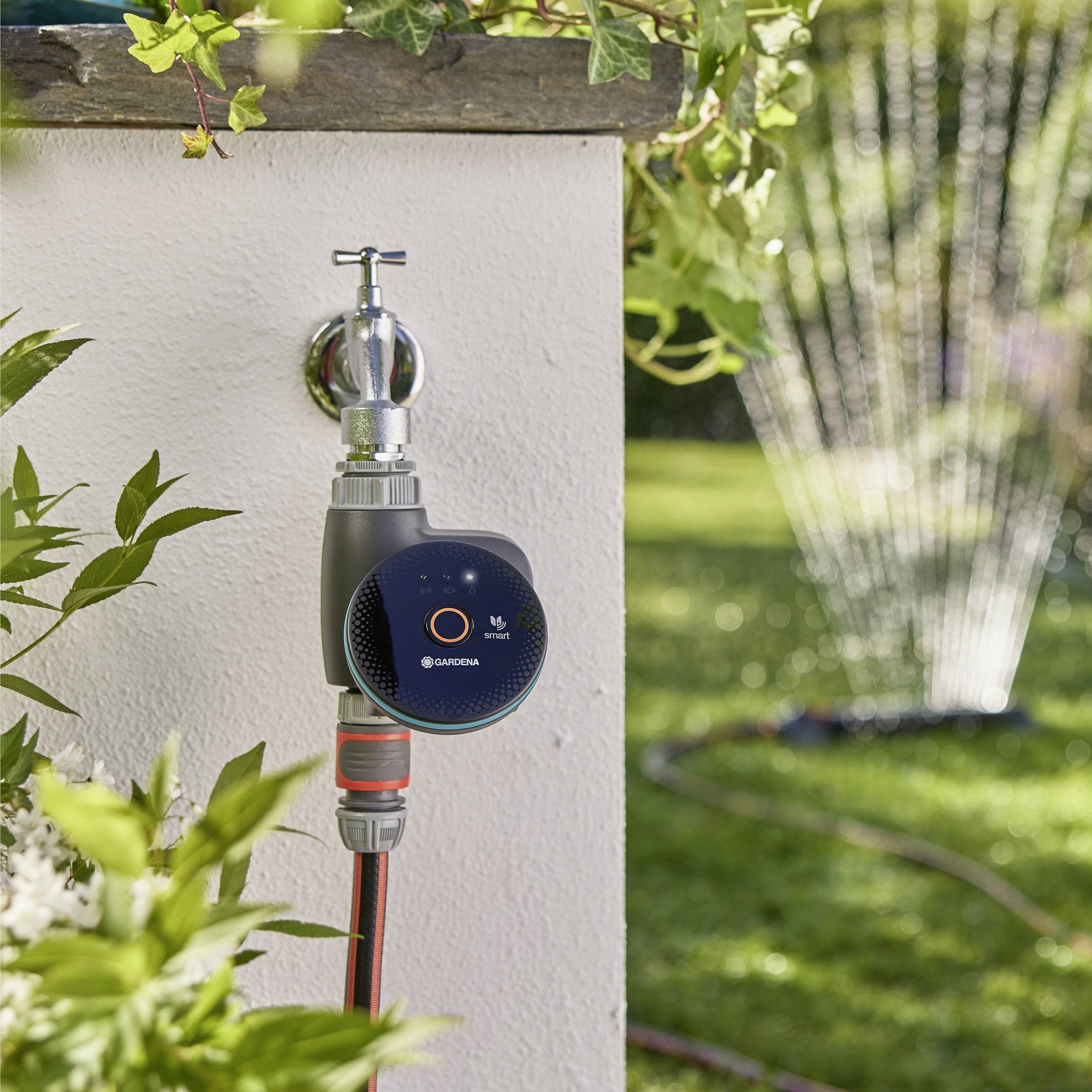 Een tuinmuur-gemonteerde watertimer bestuurt een slang, geplaatst in een zonnige tuin met groen. Een sproeier besproeit het gras op de achtergrond.