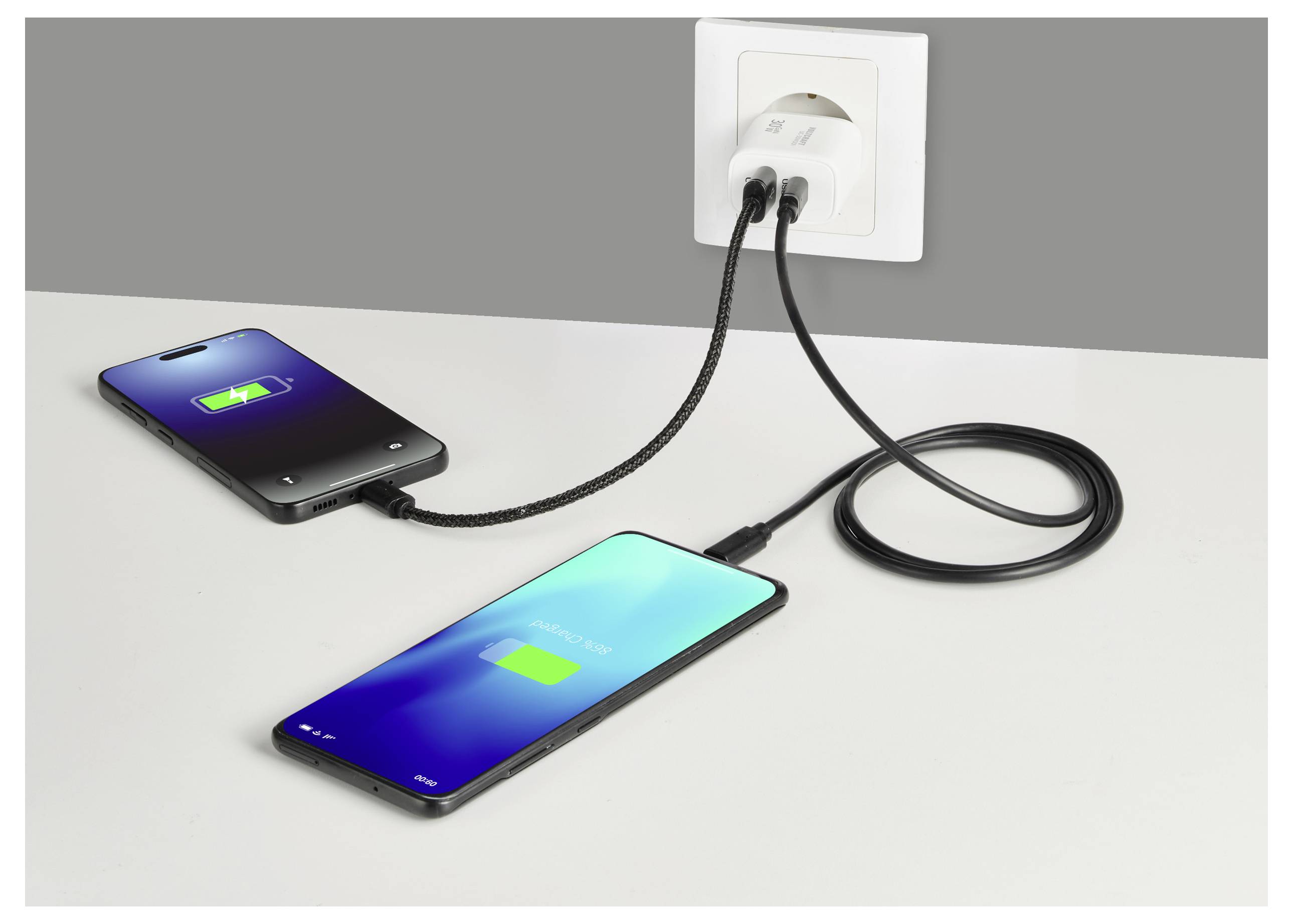 Twee smartphones laden via een dual-poort wandcontactdoos. Beide schermen tonen batterijlaadpictogrammen, wat actieve stroomoverdracht aangeeft.