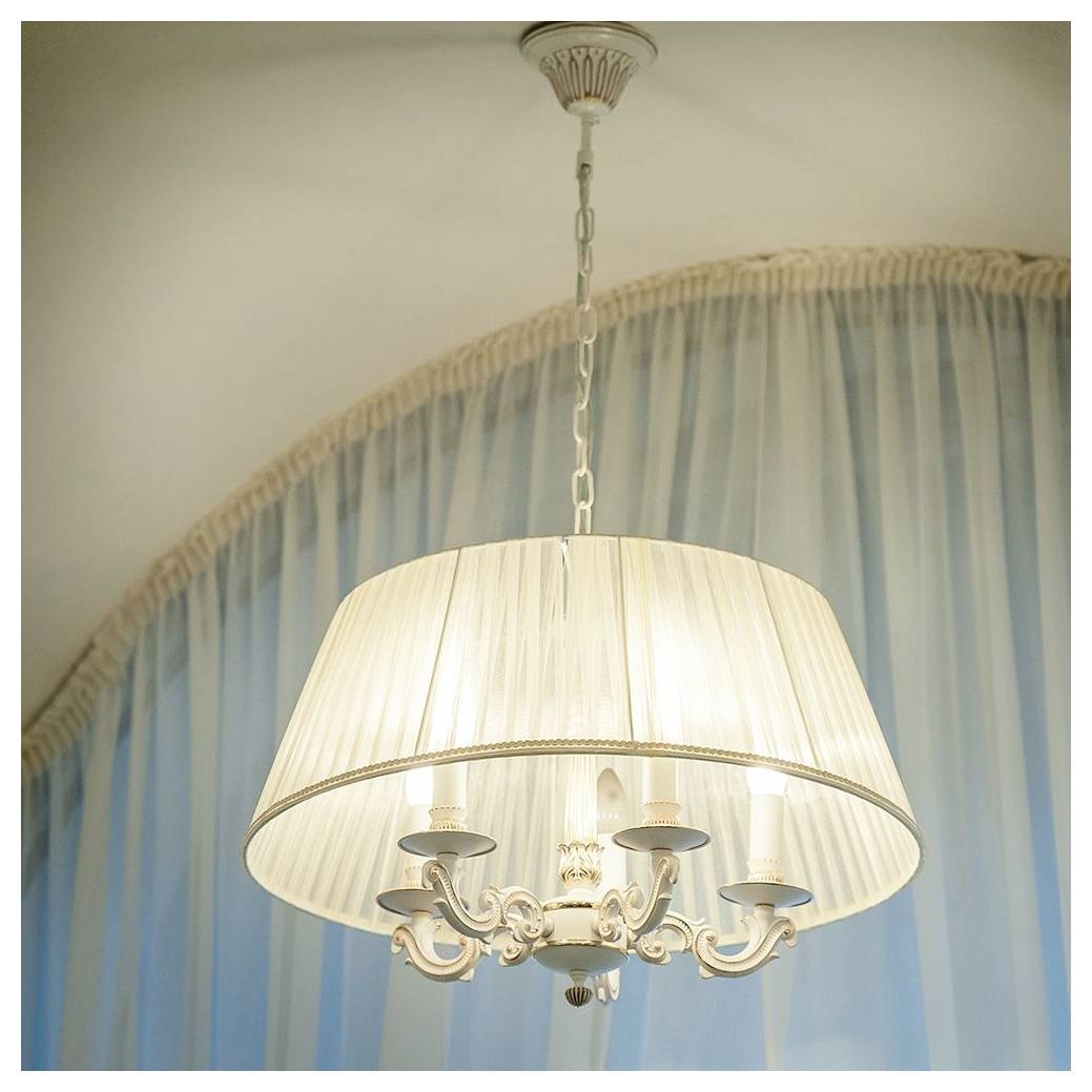 Een decoratieve kroonluchter met vijf kaarsachtige lampen en een transparante lampenkap, hangend tegen een achtergrond van zachte blauwe gordijnen.