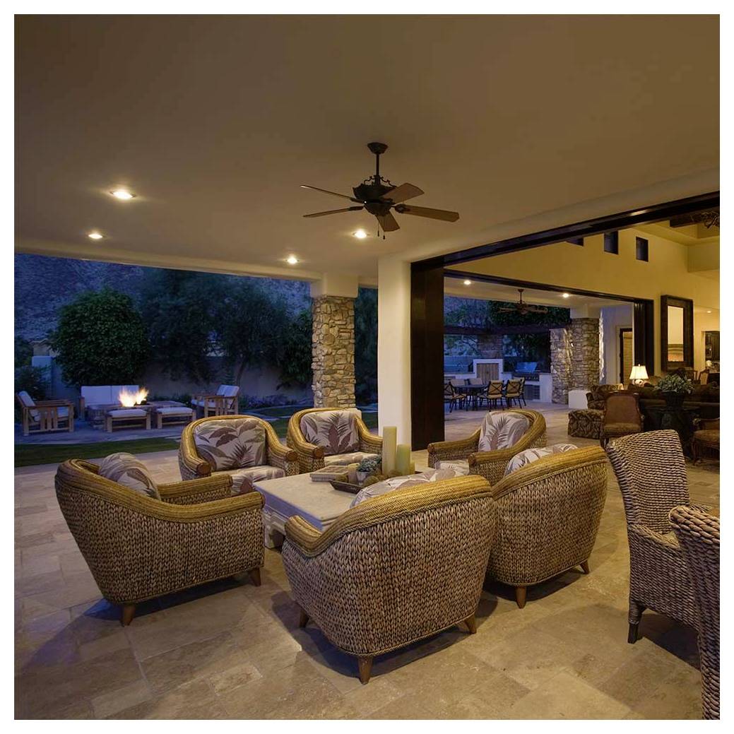 Buitenterras met rieten meubelset onder een verlichte plafondventilator. Stenen zuilen en sfeervol verlichtingsarmaturen creëren een gezellige avondsfeer.