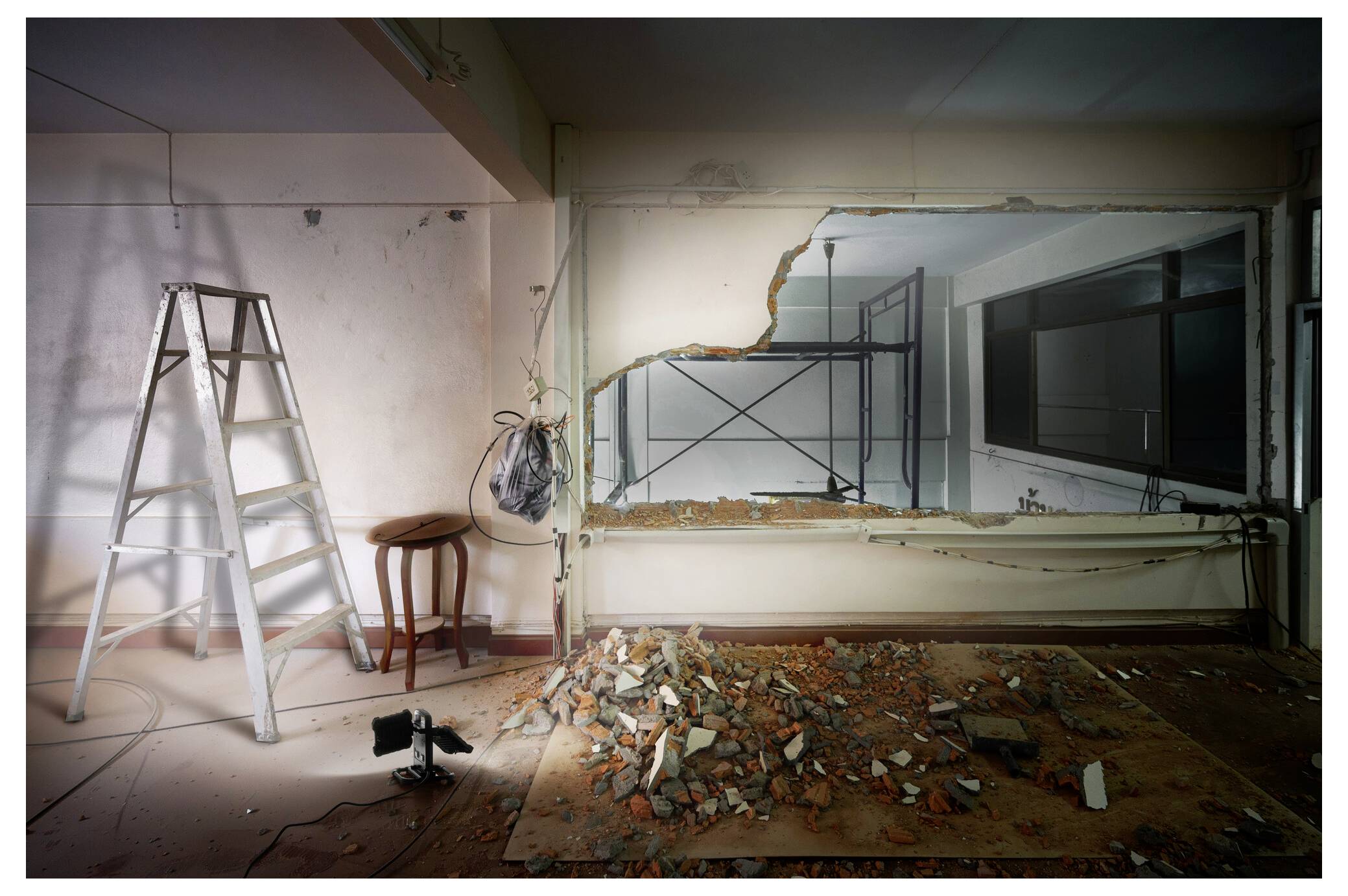 Een gedeeltelijk gesloopte muur onthult een zicht op een andere kamer. Puin is verspreid over de vloer. Een ladder en een kruk staan in de buurt.