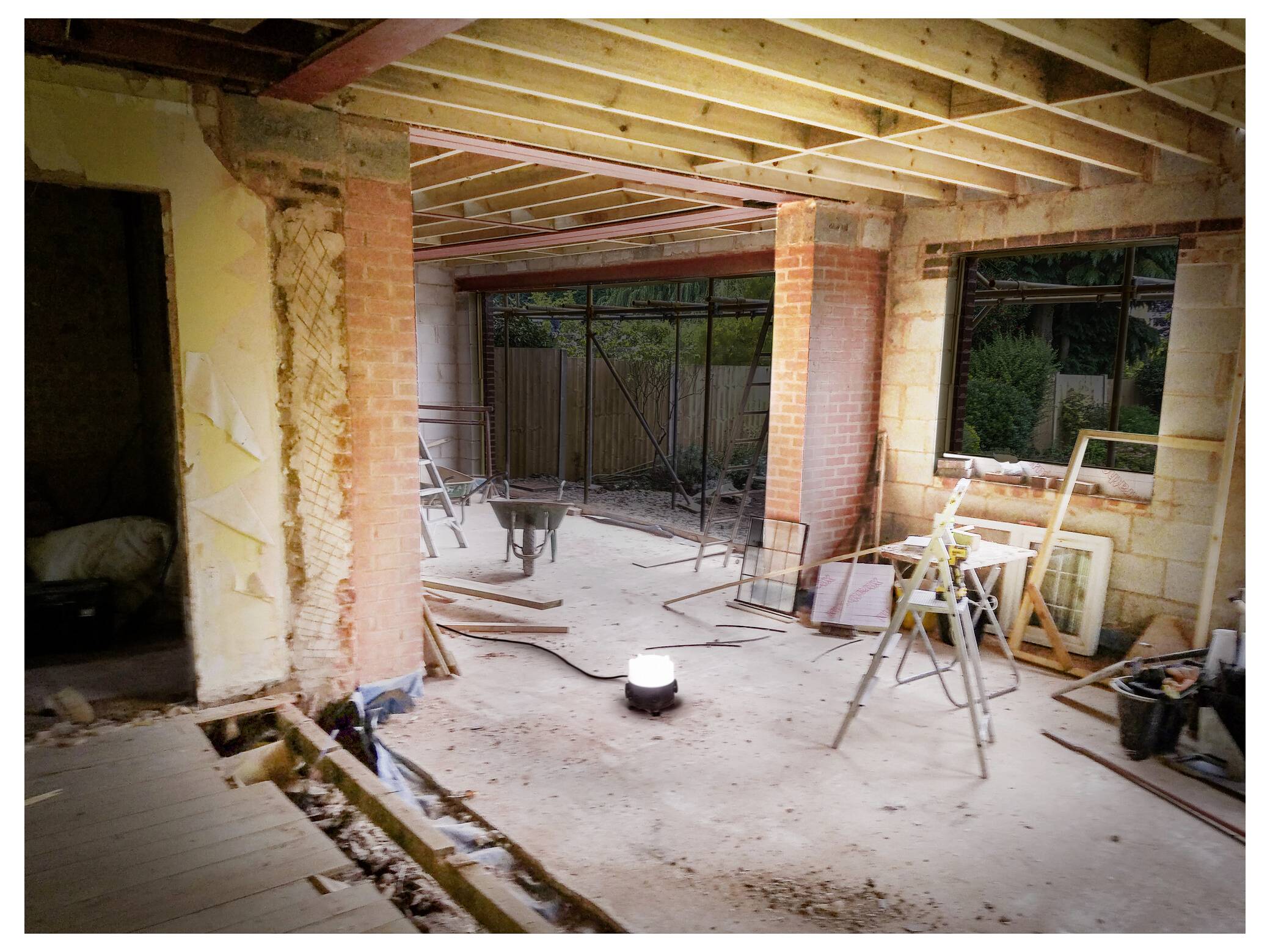 Een gedeeltelijk gerenoveerde ruimte met blootgestelde bakstenen muren en balken, bouwgereedschap en een schraag, bekeken vanuit het perspectief van een woningrenovatieproject.