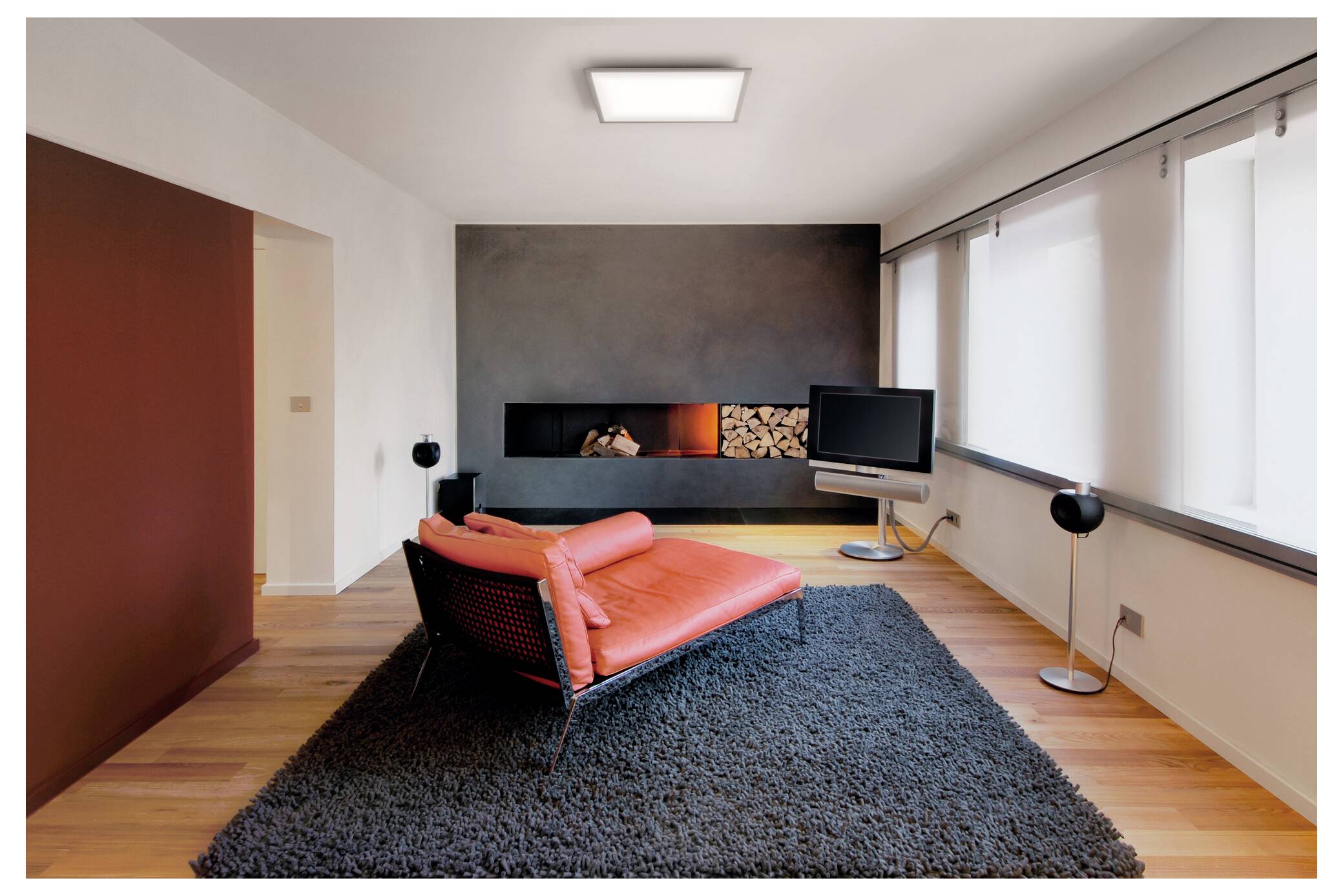 Een moderne woonkamer met een rode chaise longue op een donker vloerkleed, een flatscreen-tv en een muur met een ingebouwde haard met gestapelde houtblokken.