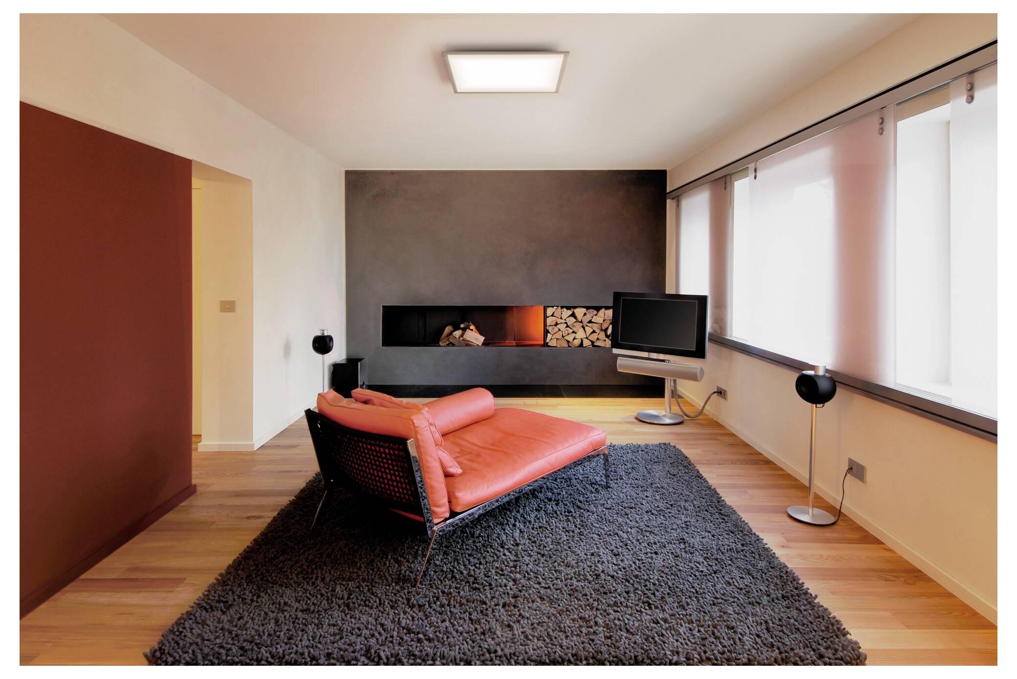 Een moderne woonkamer met een rode chaise longue op een grijze vloerkleed, een aan de muur bevestigde tv, en een open haard met gestapelde houtblokken, onder zachte verlichting.