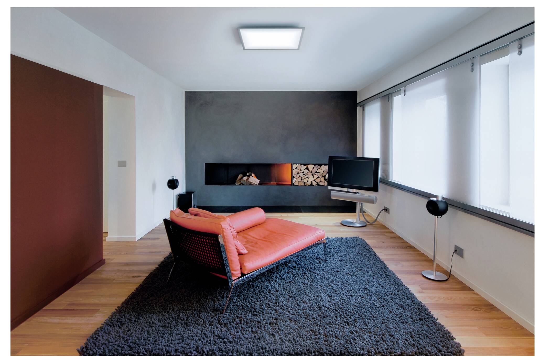 Een moderne woonkamer met een felrode chaise longue op een donker vloerkleed, grote ramen, een flatscreen-tv aan de rechterkant en een open haard aan de linkerkant.