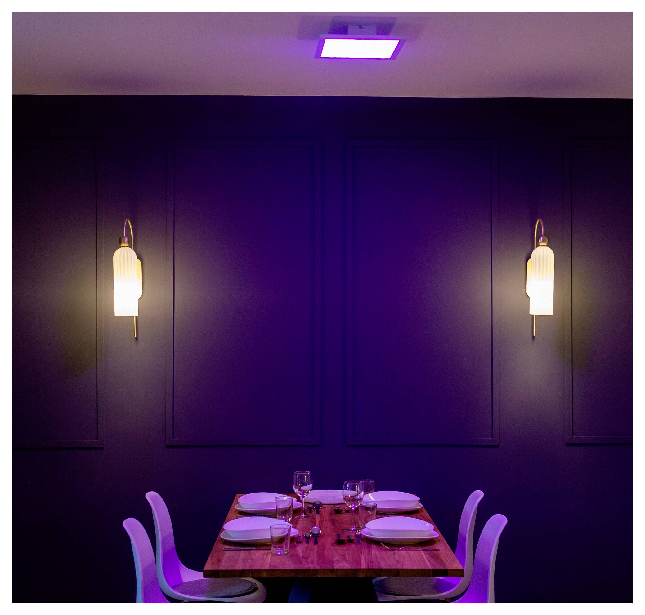 Een schaars verlichte eetkamer met een houten tafel gedekt voor vier personen, met witte stoelen, borden, glazen en twee wandlampen tegen een donkere achtergrond.