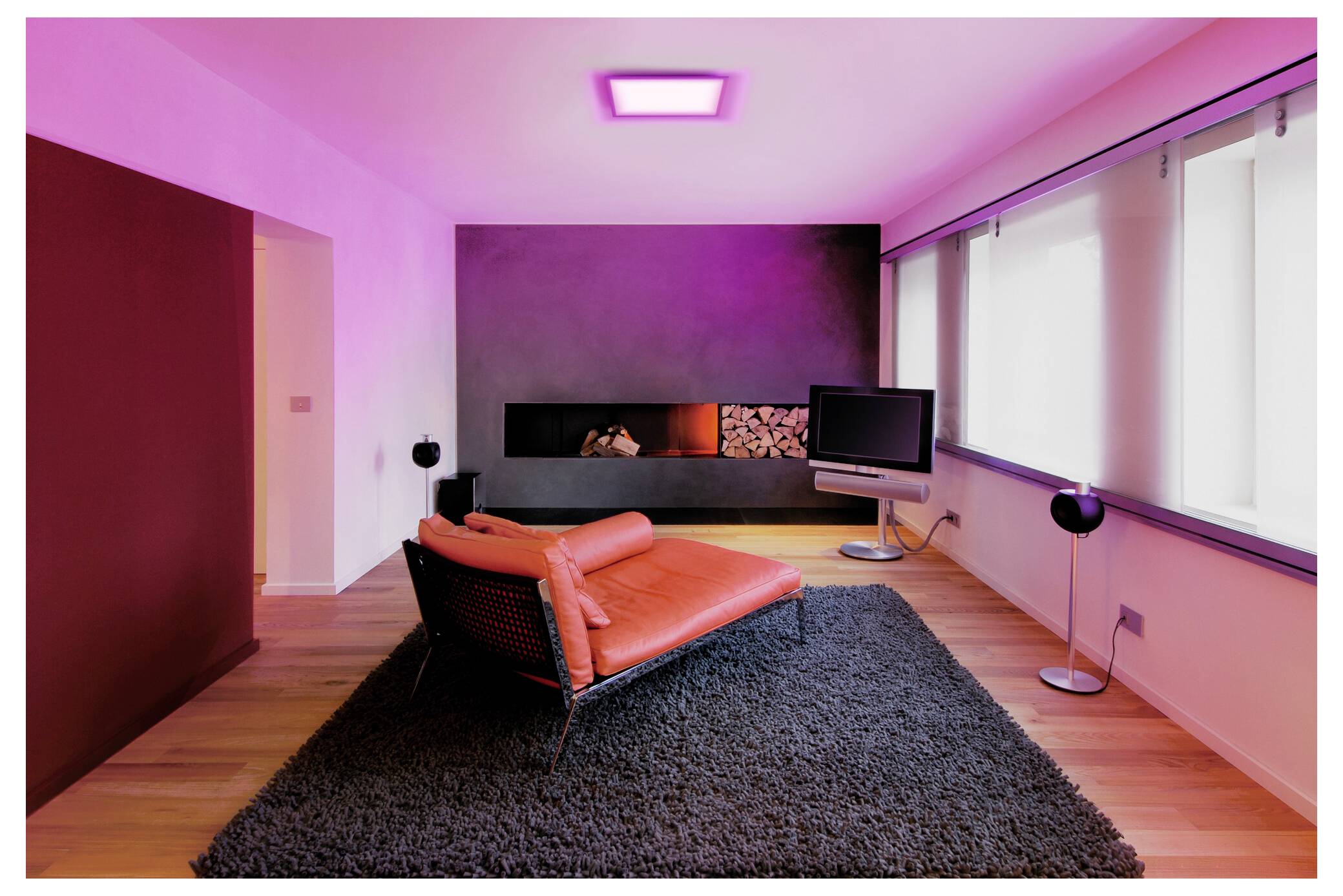 Een moderne woonkamer met een rode chaise longue, een zwart tapijt, een aan de muur bevestigde tv en een open haard. De ruimte wordt verlicht met roze verlichting.