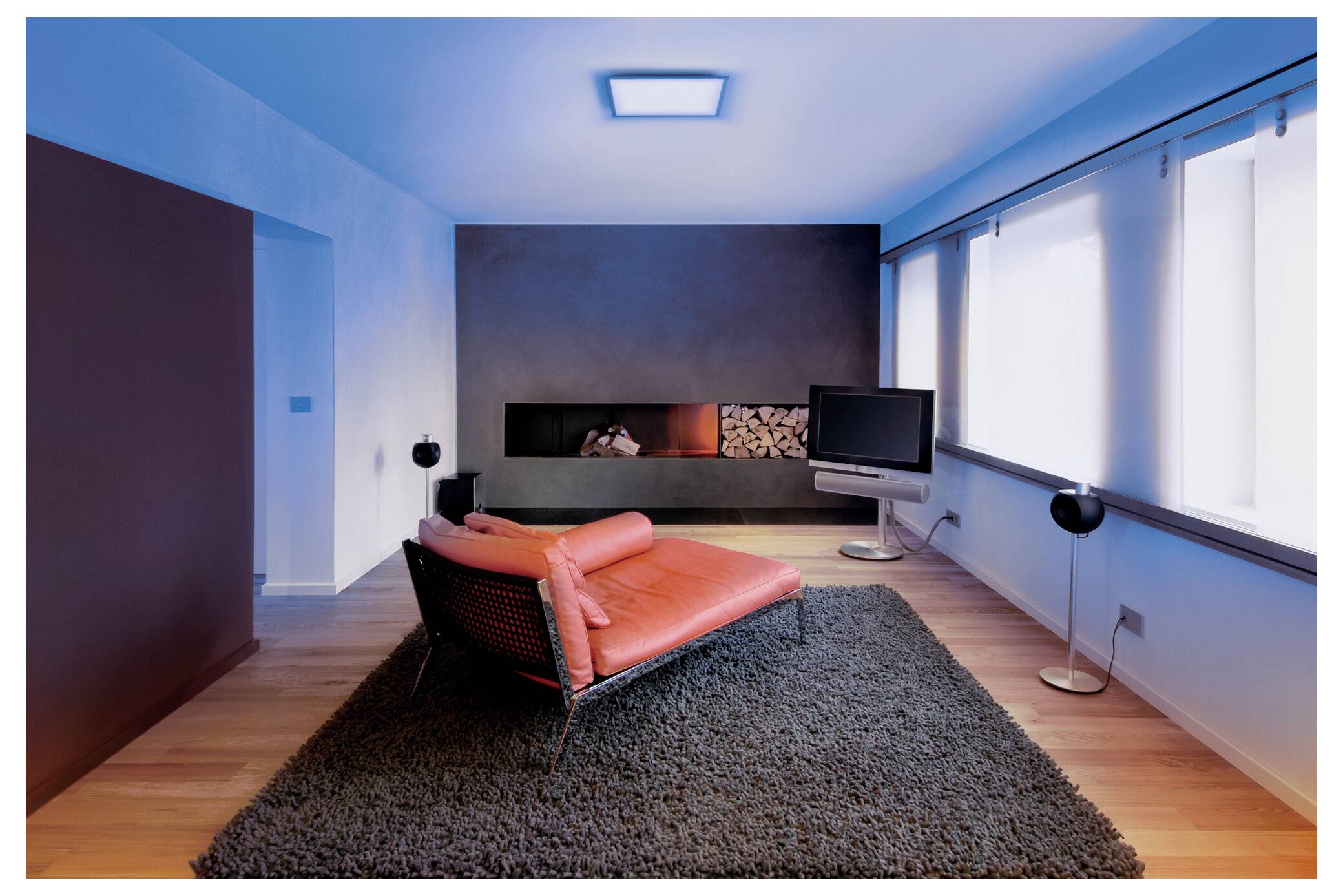 Een moderne woonkamer met een rode loungestoel op een grijze vloerkleed, een flatscreen-tv en een openhaard met houtblokken. Grote ramen laten natuurlijk licht binnen.