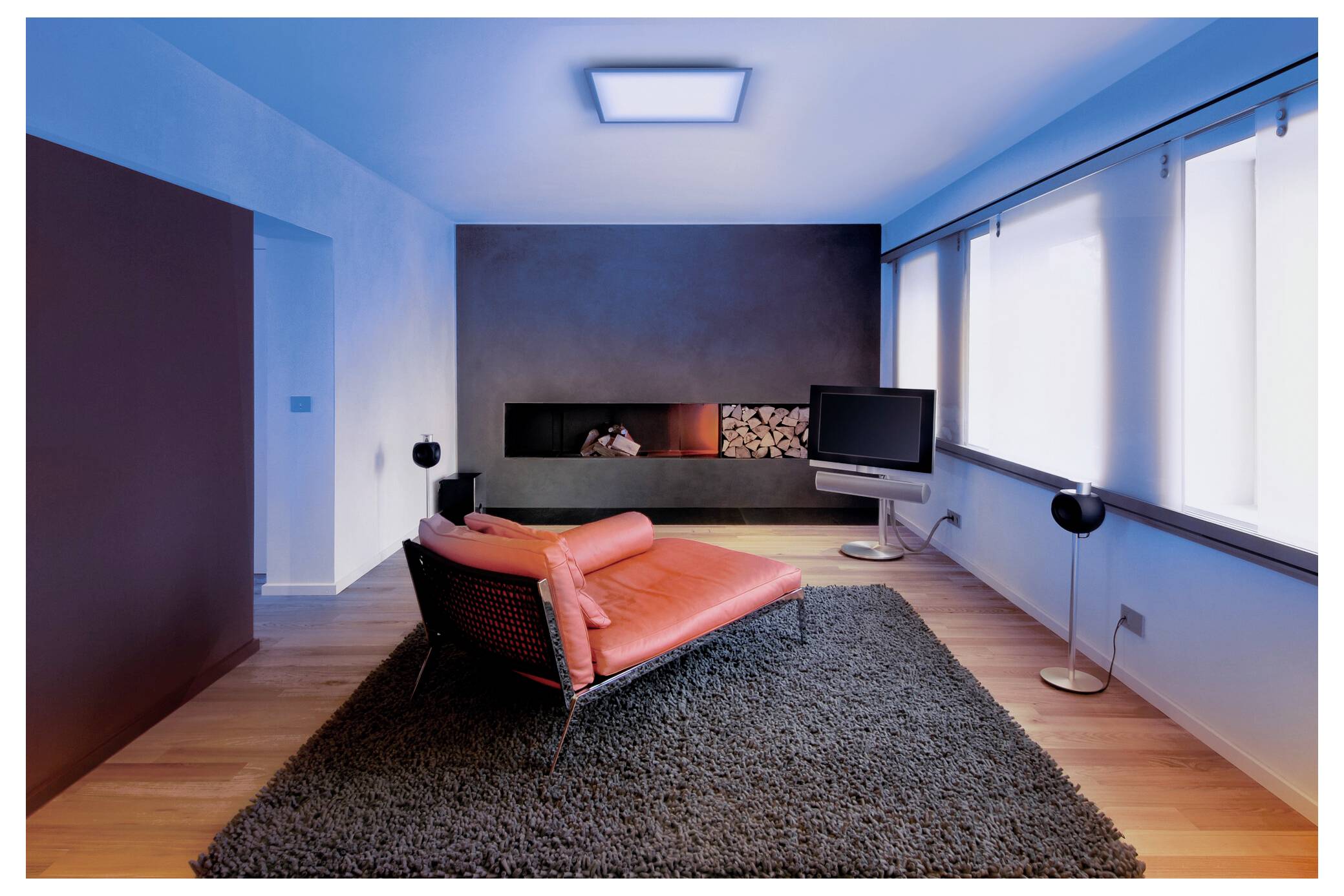 Een moderne woonkamer met een rode chaise longue op een donker tapijt, een aan de muur bevestigde open haard, een flatscreen-tv en grote ramen aan de rechterkant.