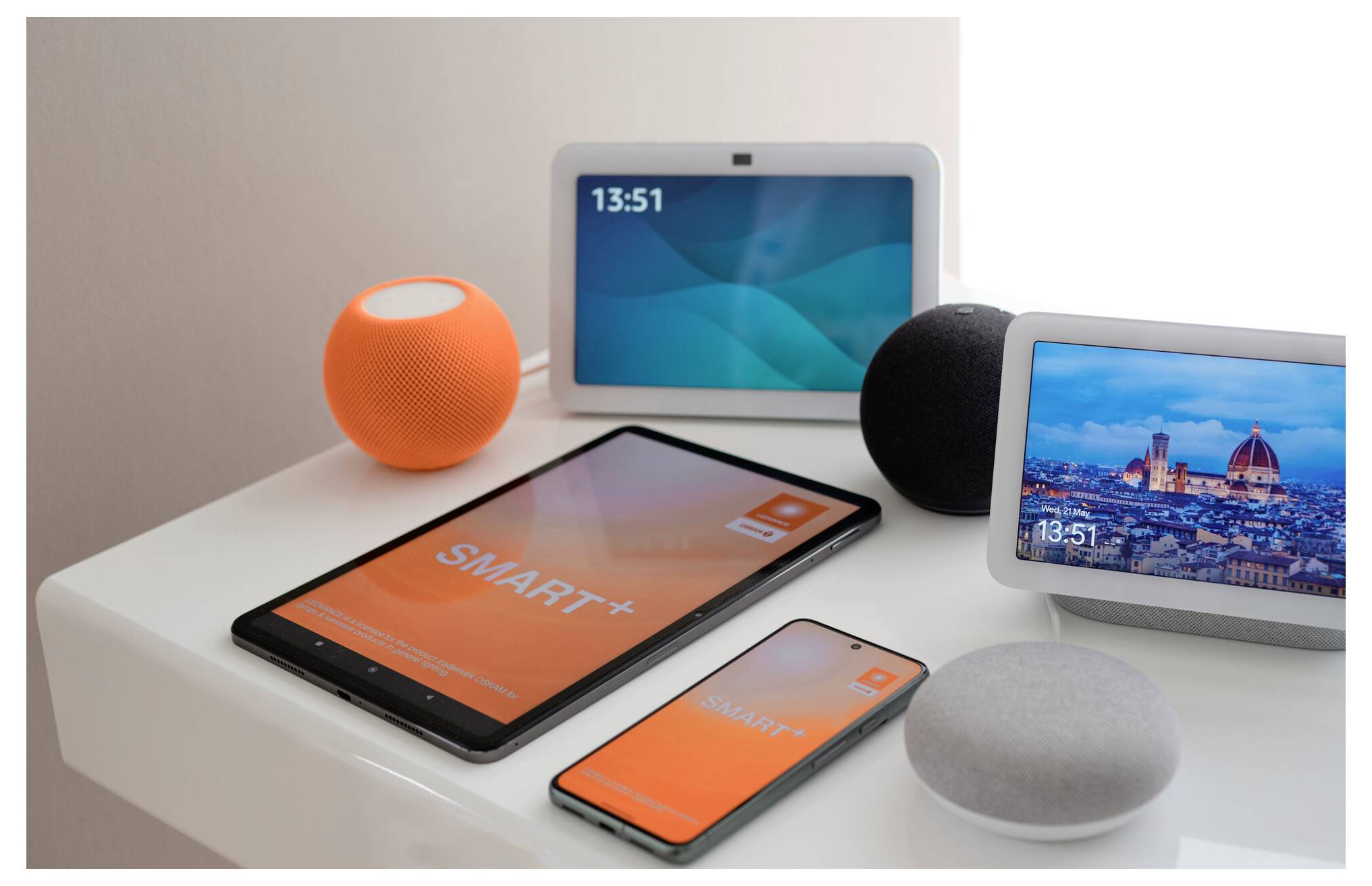 Verschillende slimme apparaten, waaronder tablets, een smartphone, smart speakers en displays, zijn op een witte tafel geplaatst, wat wijst op een technologisch thema.