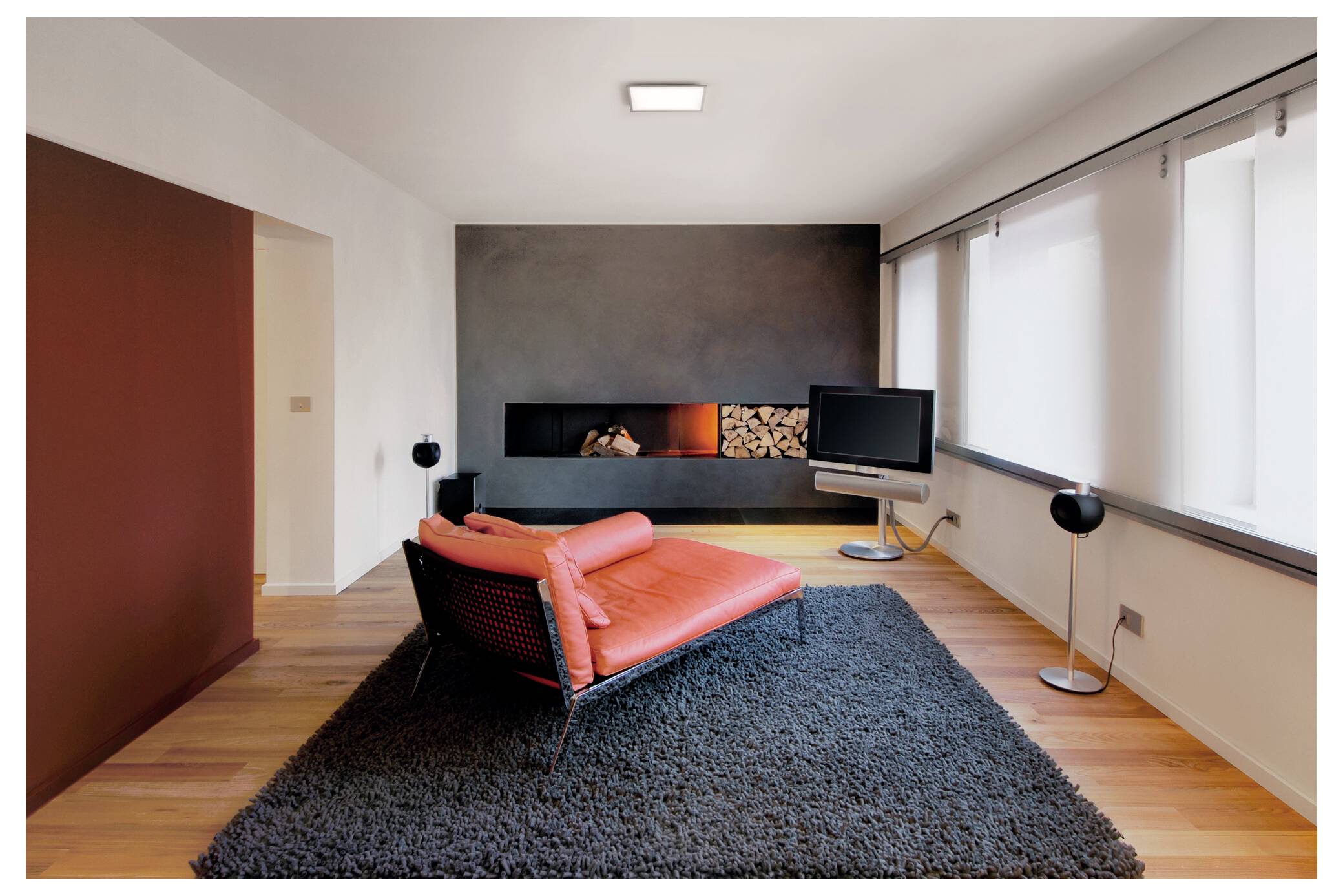 Een moderne woonkamer met een rode chaise longue op een grijze vloerkleed, een zwarte accent muur met opgestapeld brandhout, en een groot flatscreen-tv-toestel op een standaard.