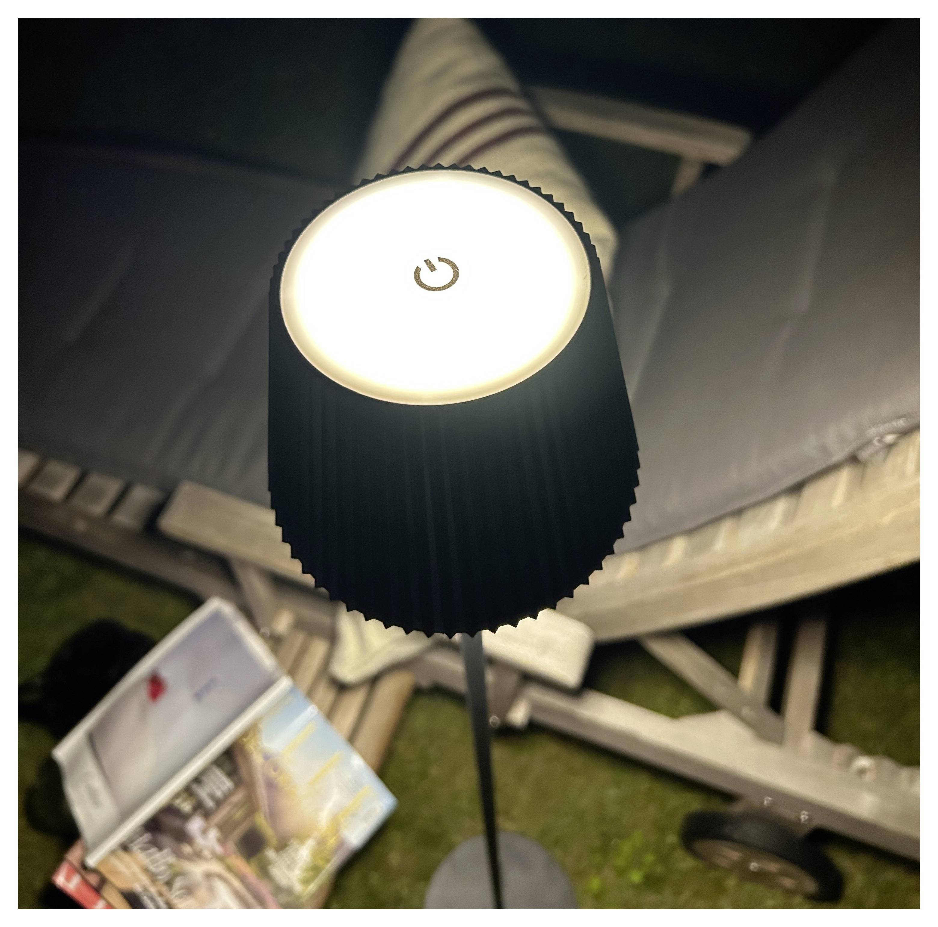 Een staande LED-lamp met een aan/uit-knop icoon bovenop, die een ruimte verlicht met tijdschriften op een stoel en een kussen, wat een gezellige leeshoek suggereert.