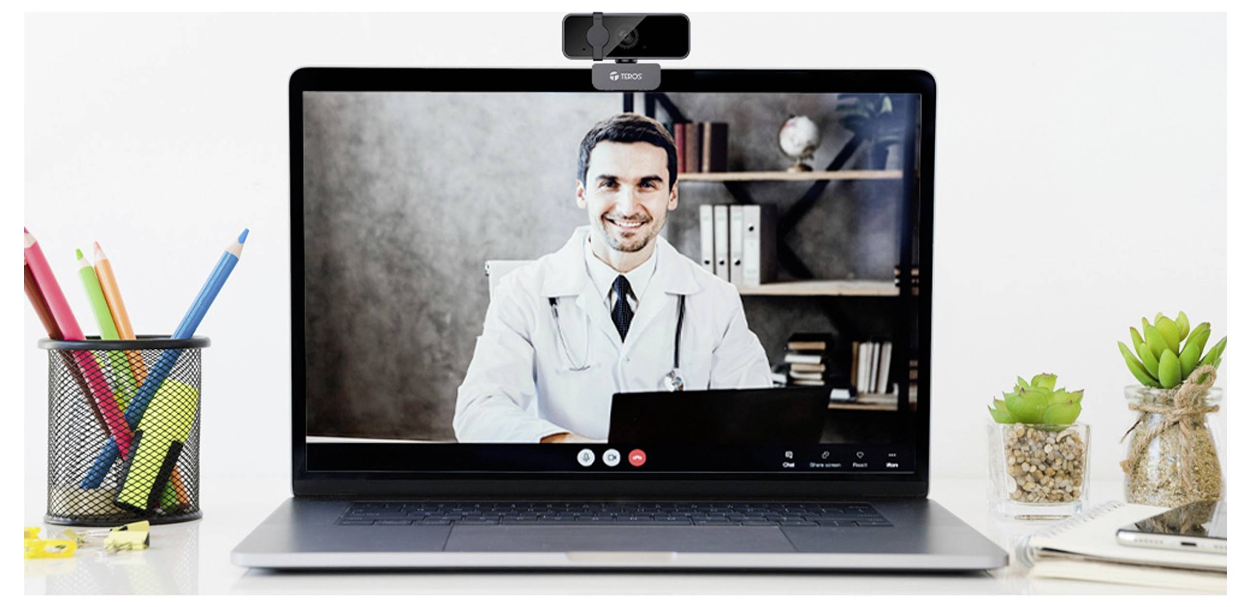 'Een arts in een witte jas videoconferentie voerend op een laptop, omringd door kantoorspullen inclusief planten en een potloodholder.'
