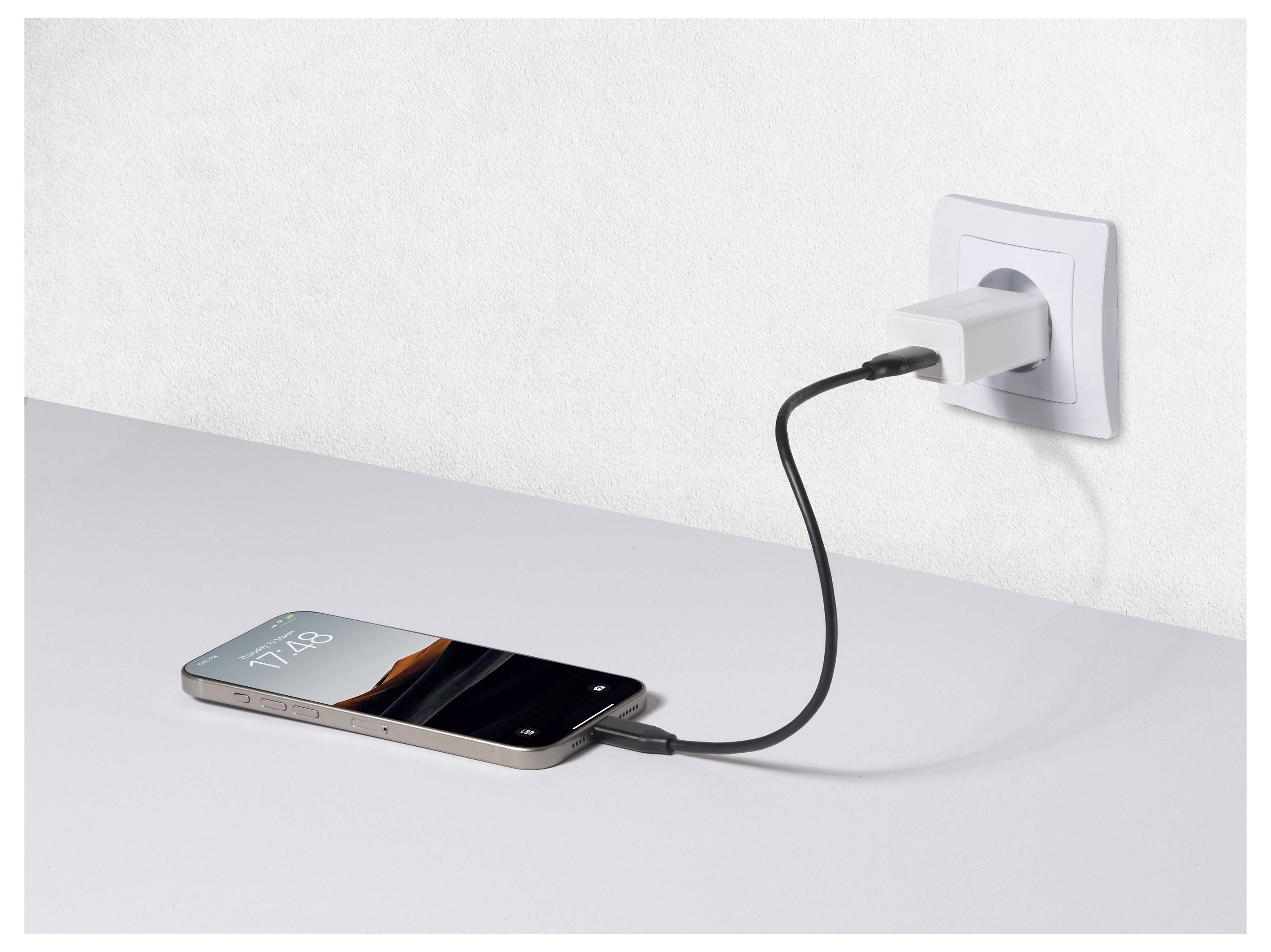 Een smartphone die oplaadt op een wit oppervlak, verbonden met een wandcontactdoos via een zwarte kabel, het scherm toont de tijd '17:25'.