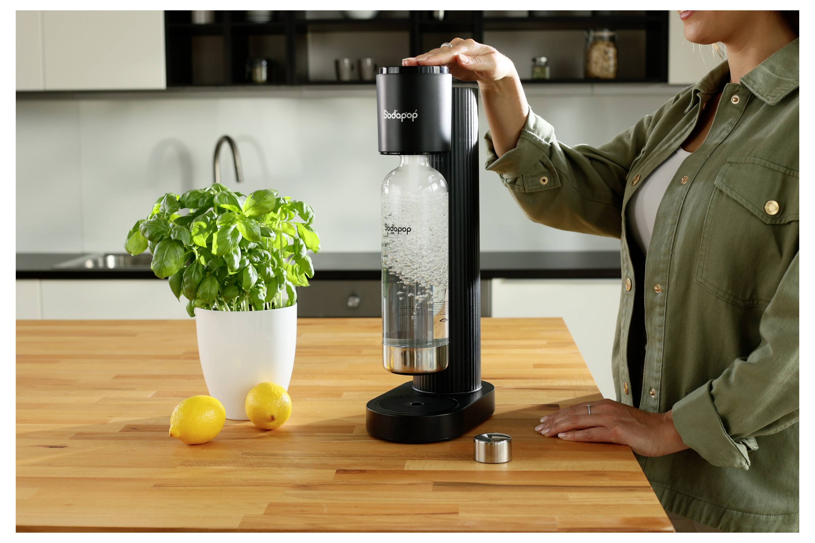 Een persoon bedient een bruiswatermachine op een keukenwerkblad naast een potplant met basilicum en twee citroenen. Het apparaat is water aan het koolzuren in een fles.