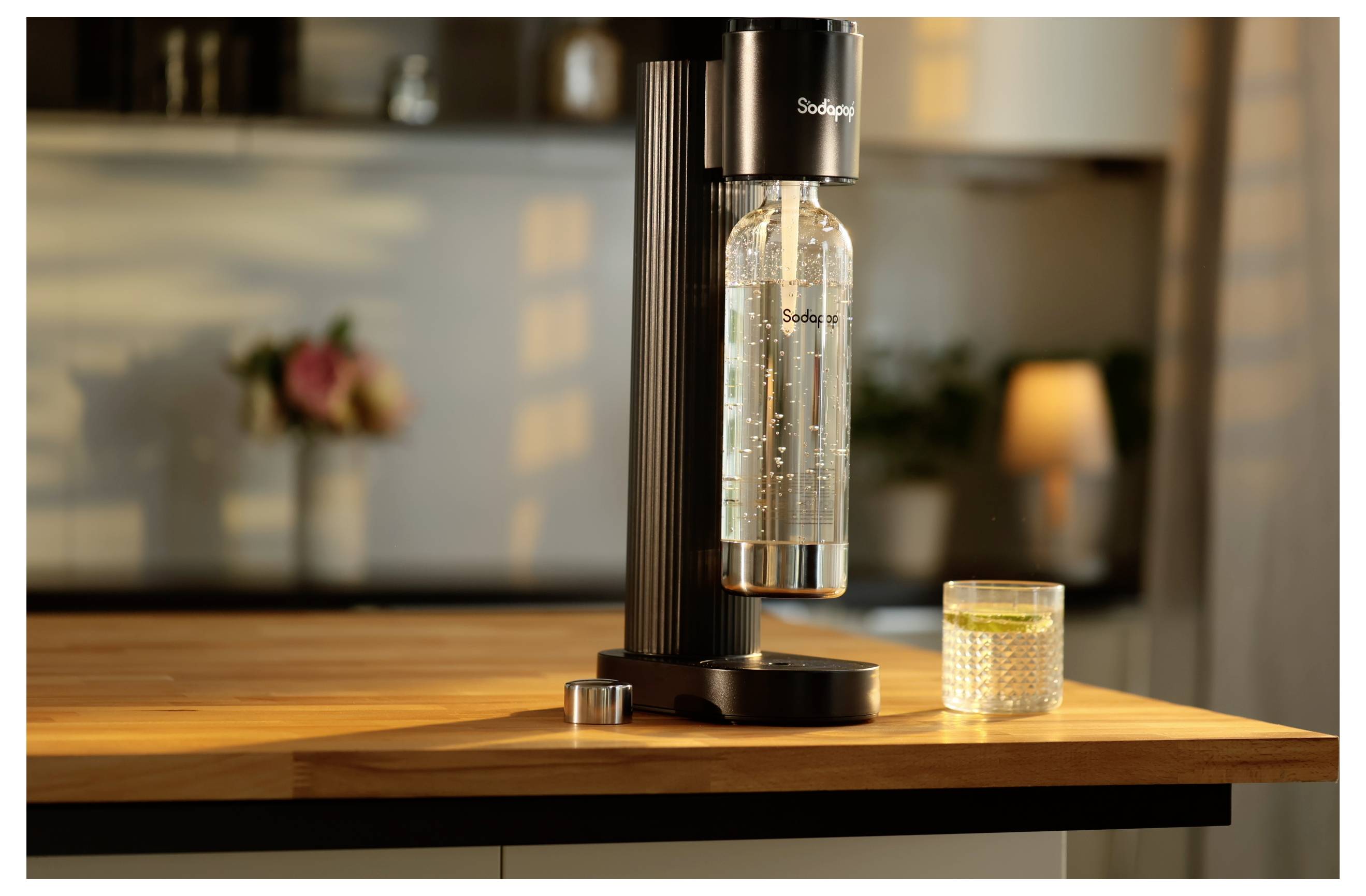 Een zwarte sodamaker op een aanrecht met een transparante fles eraan bevestigd, naast een gevuld getextureerd glas, in een warmverlichte keukenomgeving.