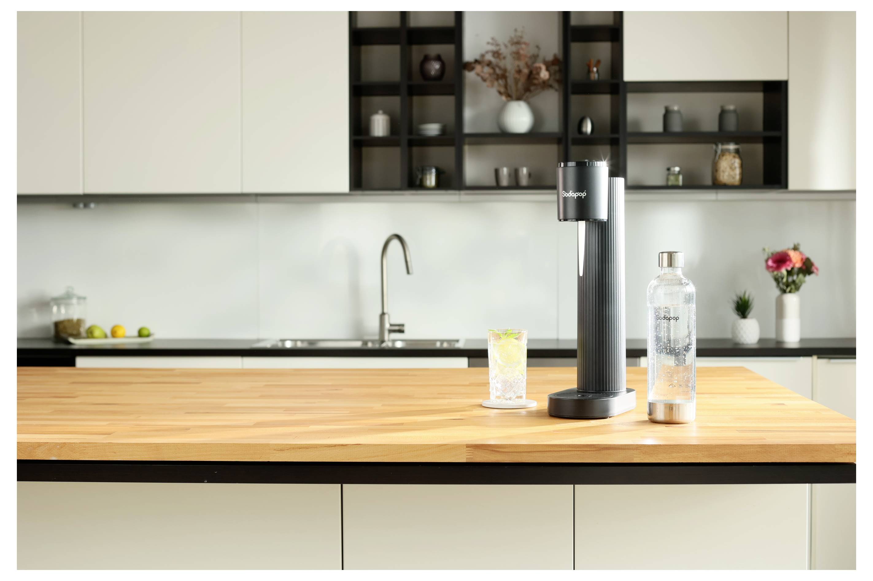 Een moderne keukentoonbank met een bruiswater-apparaat, een glas citroenwater en een fles. Planken op de achtergrond met decoratieve items.