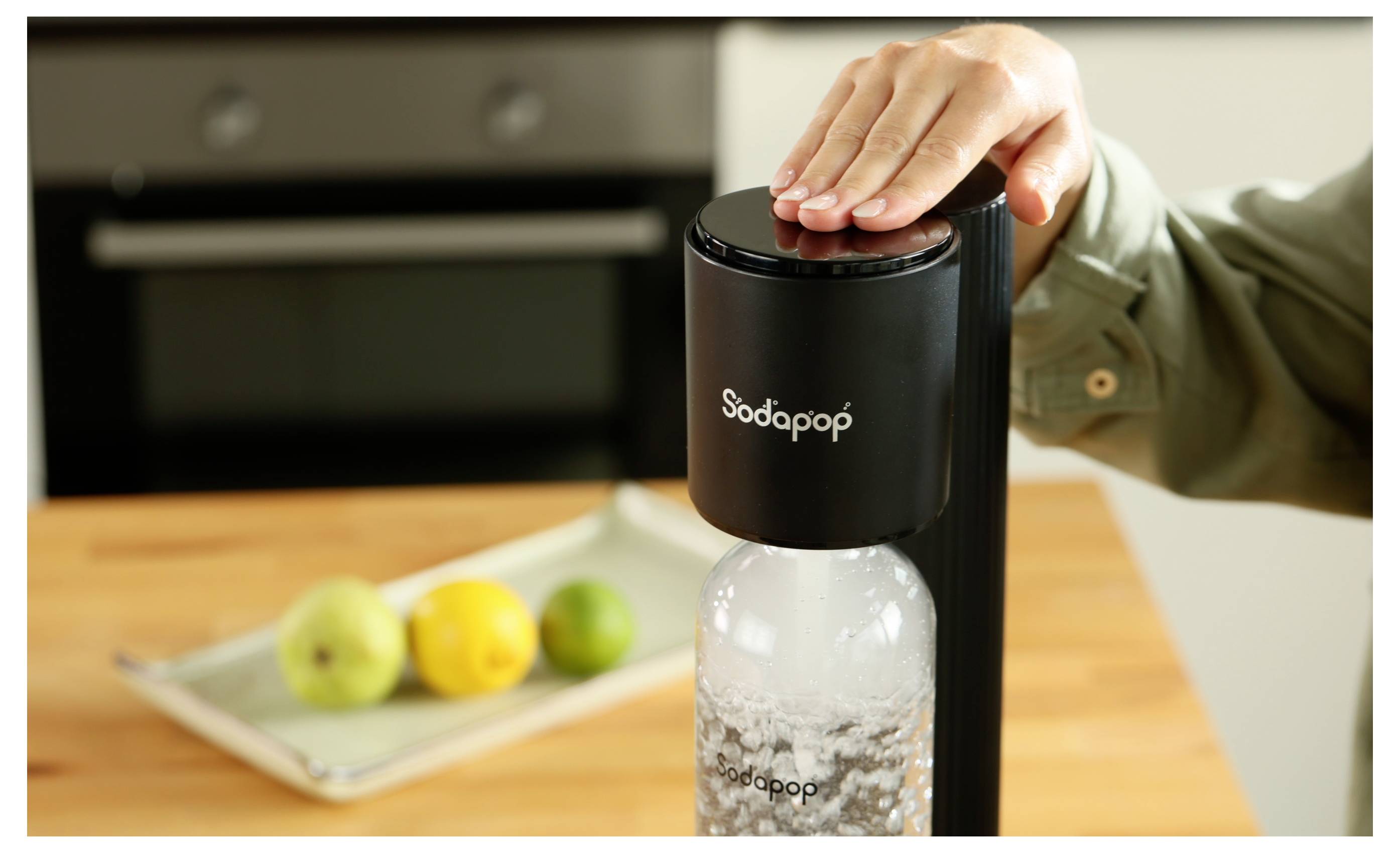 Een hand drukt op de bovenkant van een Sodapop-machine, waardoor water wordt gecarbonateerd in een fles. Een dienblad met fruit is zichtbaar op de achtergrond op een aanrecht.