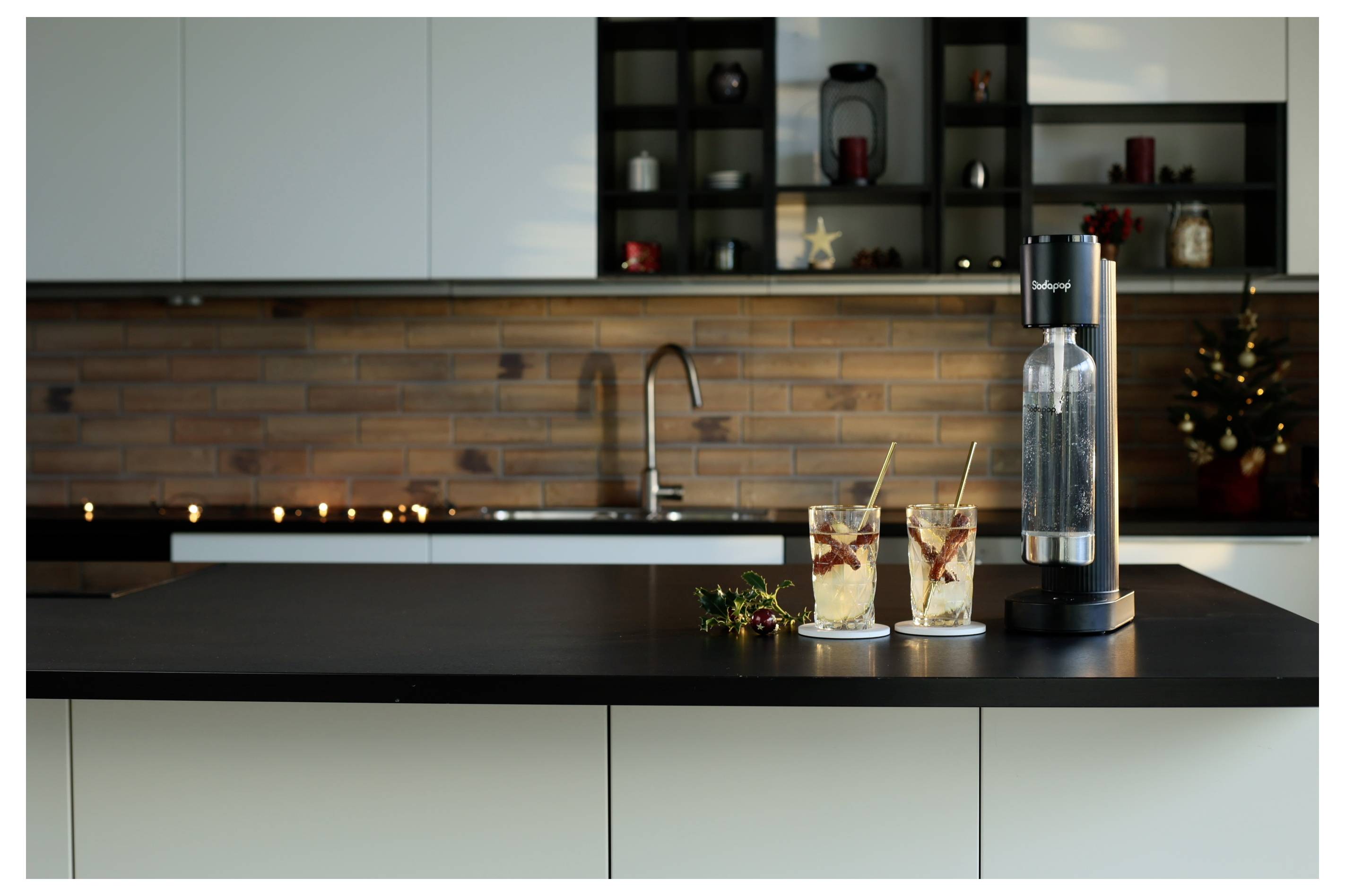 Moderne keuken met zwart aanrechtblad, een sodamaker en twee glazen ijskoffie met rietjes. Bakstenen achterwand en zacht licht.