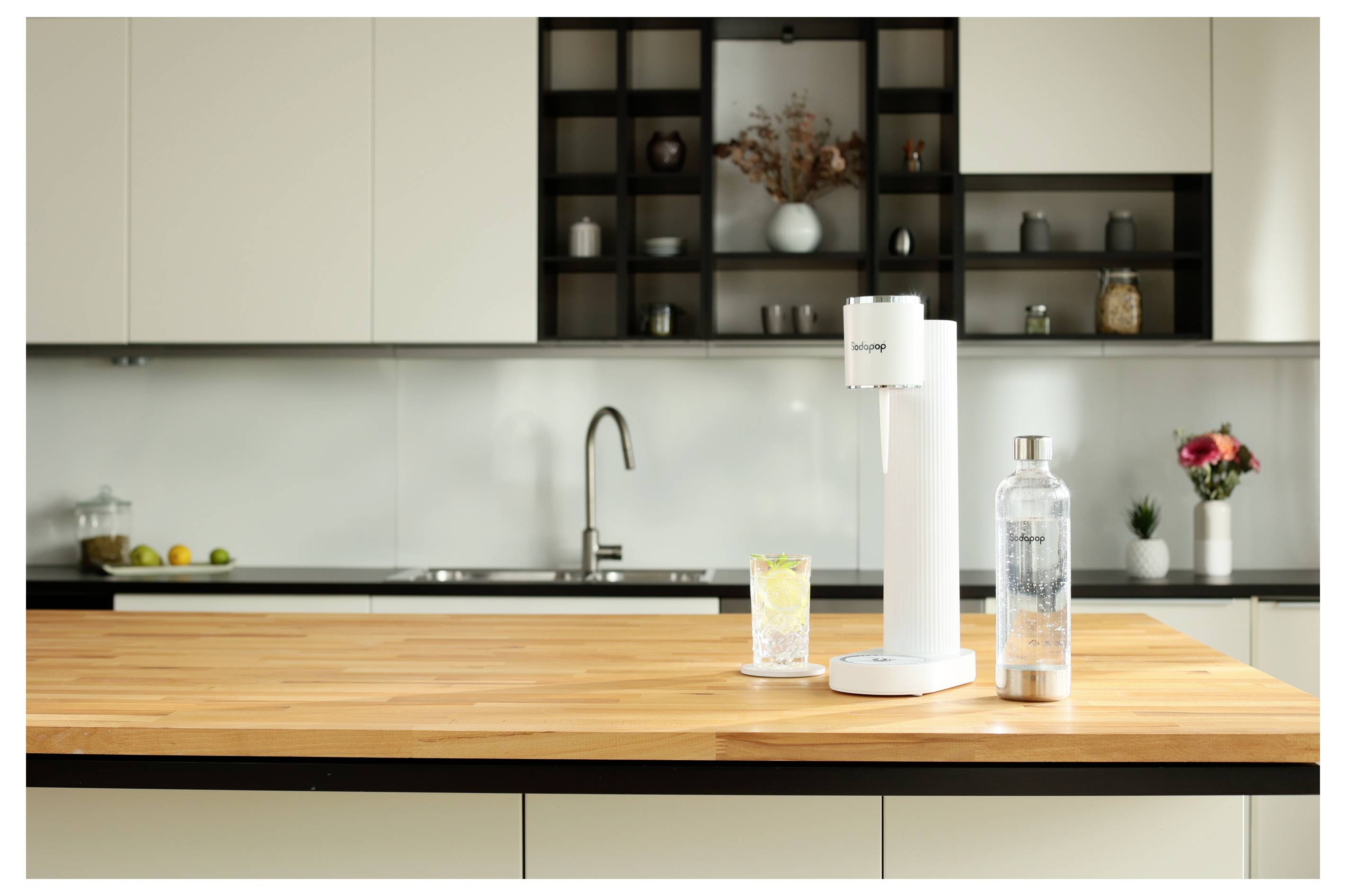 Een witte keukenwerkblad met een drankkoolzuurapparaat, een gevuld glas en een fles in focus. Moderne kasten en decoratie op de achtergrond.