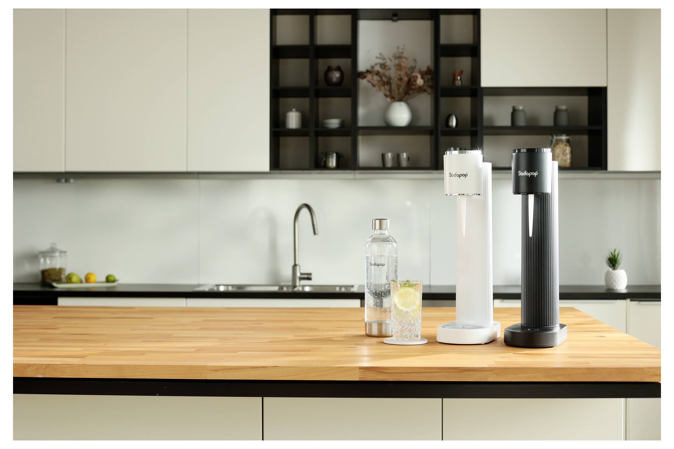 Een moderne keuken met twee bruiswaterapparaten op een houten aanrecht, vergezeld van een glazen fles en een glas water met een schijfje citroen.