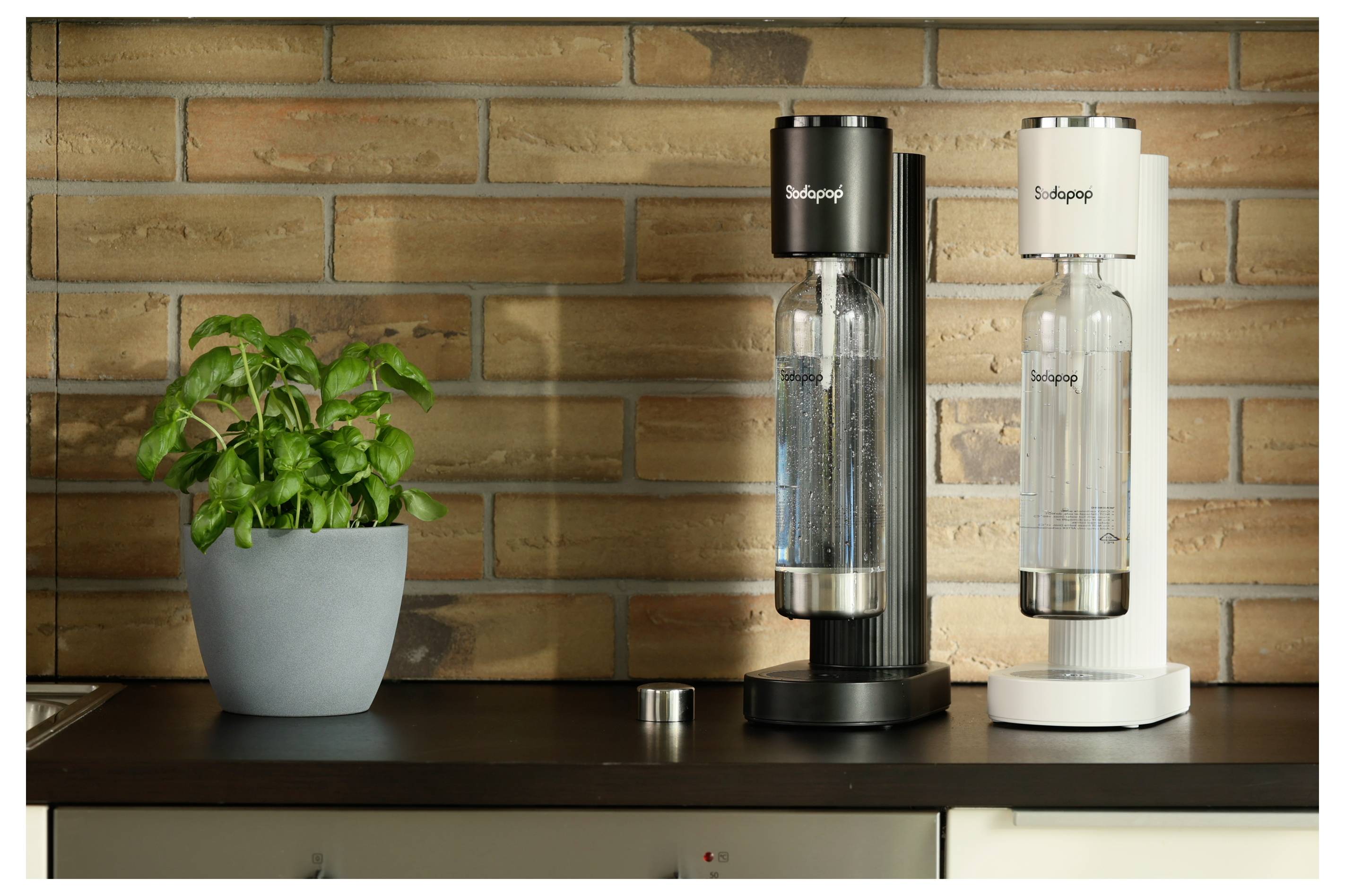 Twee koolzuurmachines, één zwart en één wit, met waterflessen op een aanrechtblad naast een potplant tegen een bakstenen muur.