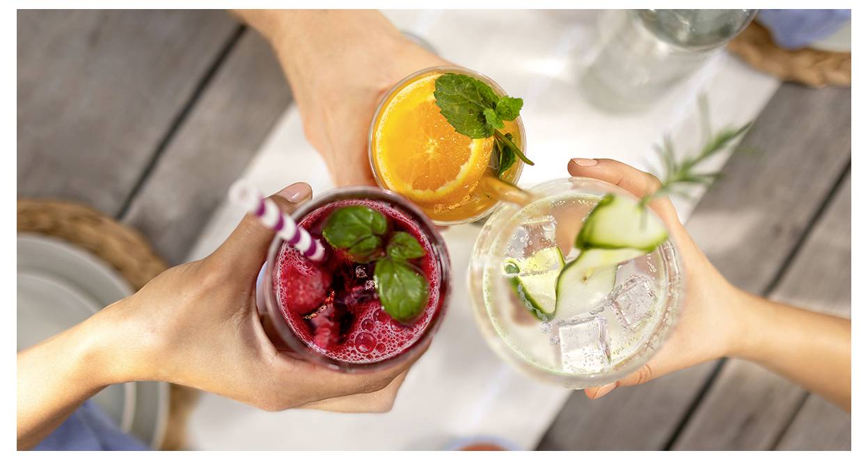Drie handen die proosten met kleurrijke drankjes: een rode met bessen, een oranje met citrus en munt, en een heldere drank met komkommer en rozemarijn.