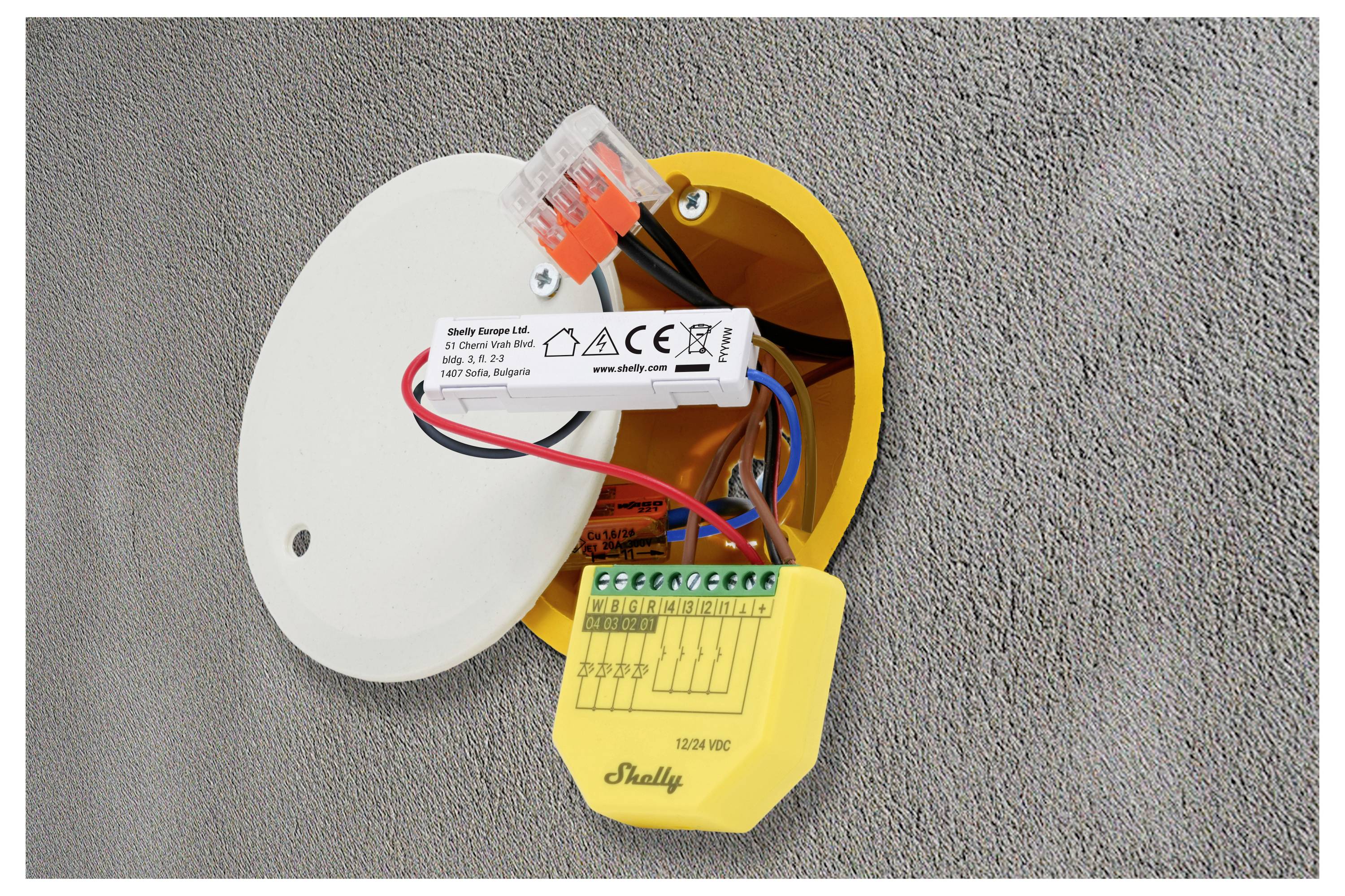 Een open elektrische verbindingsdoos in een muur met blootgestelde bedrading en componenten. De opstelling bevat een wit deksel, klemmenblokken en labels.