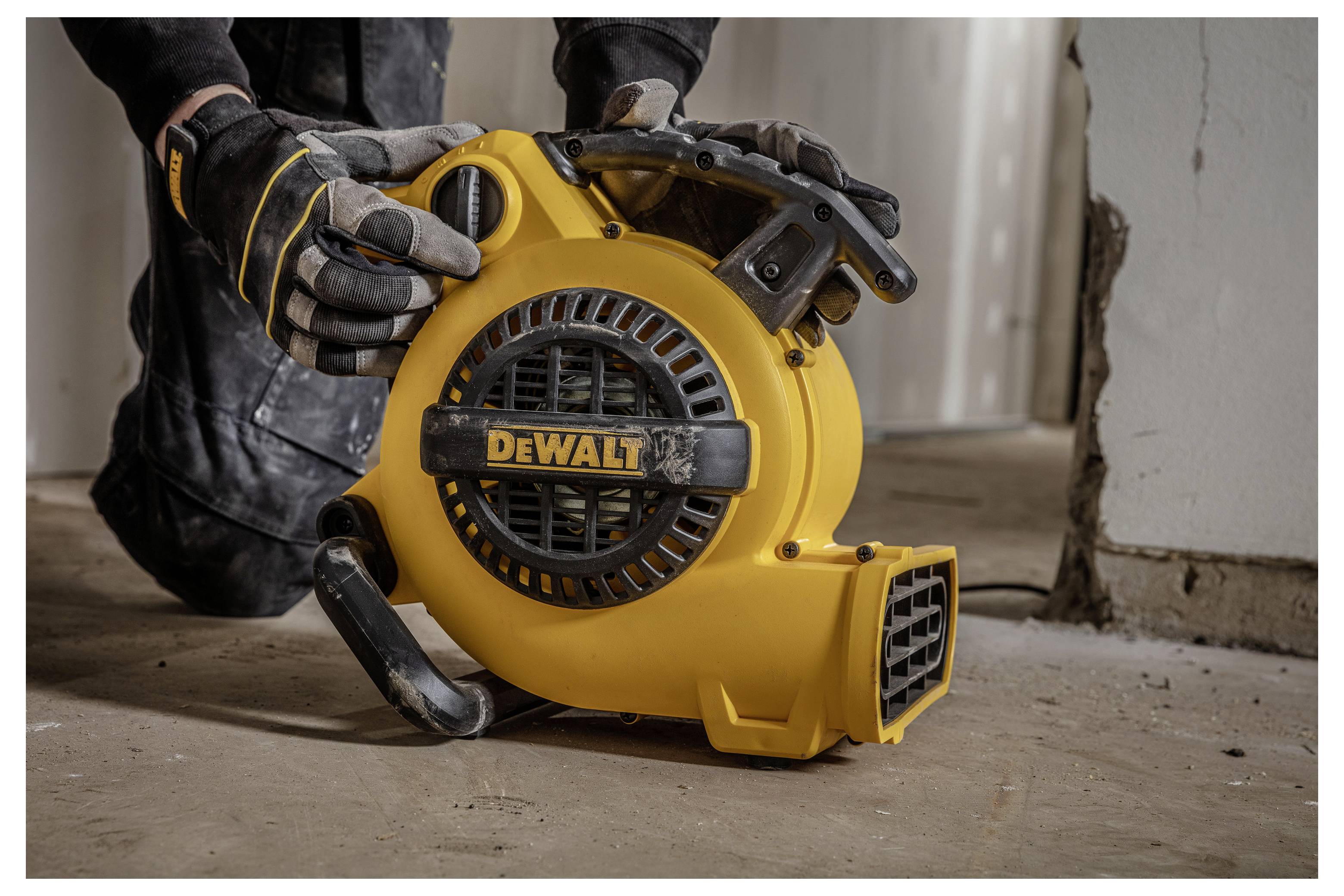 Gehandschoende werknemer die een gele DEWALT draagbare blaasmotor/ventilator op een bouwplaatsvloer vasthoudt.