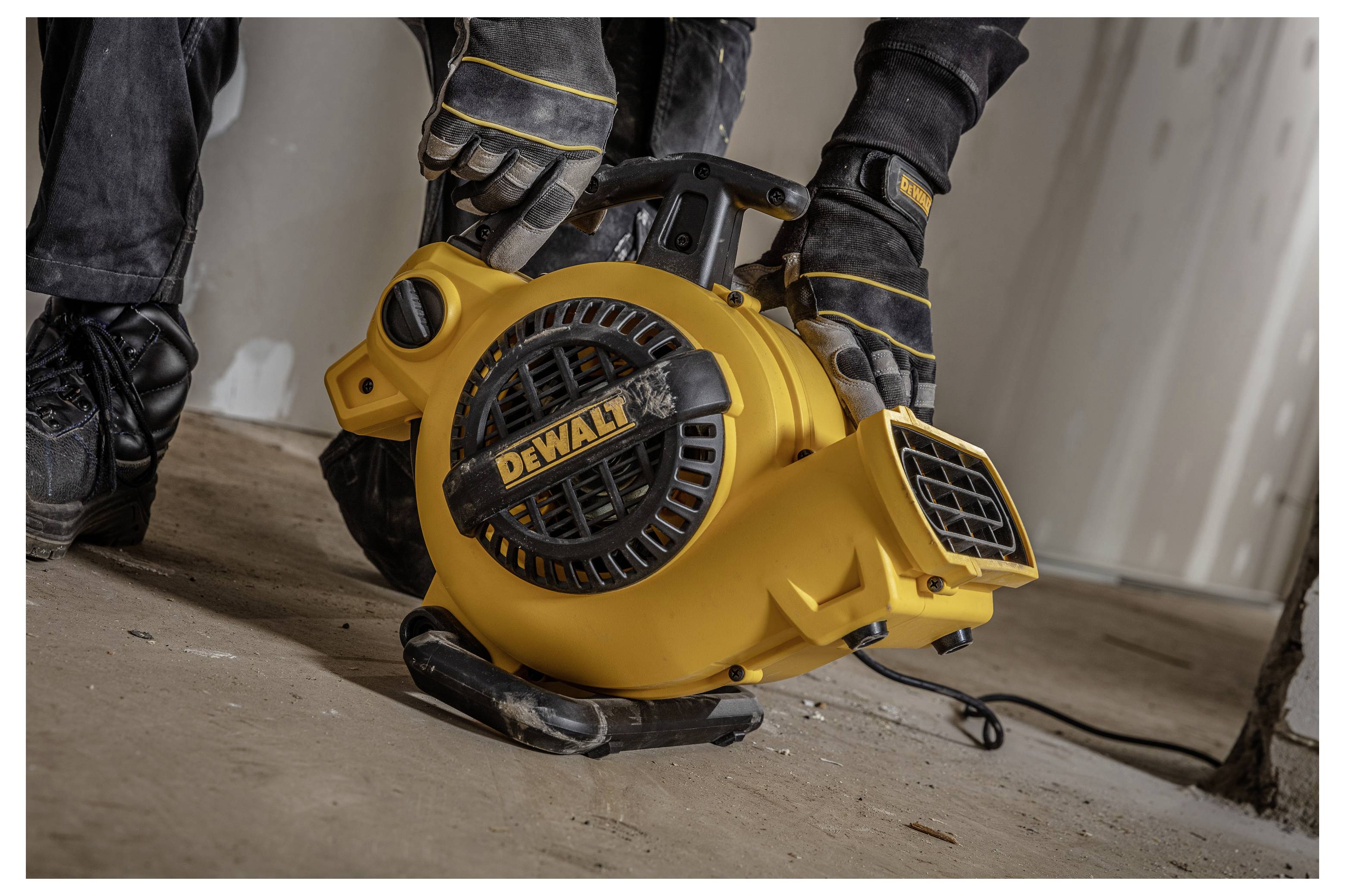 Werknemer met handschoenen houdt een gele DEWALT draagbare blaastofzuiger of ventilator vast op een stoffige vloer tijdens bouw- of schoonmaakwerk.