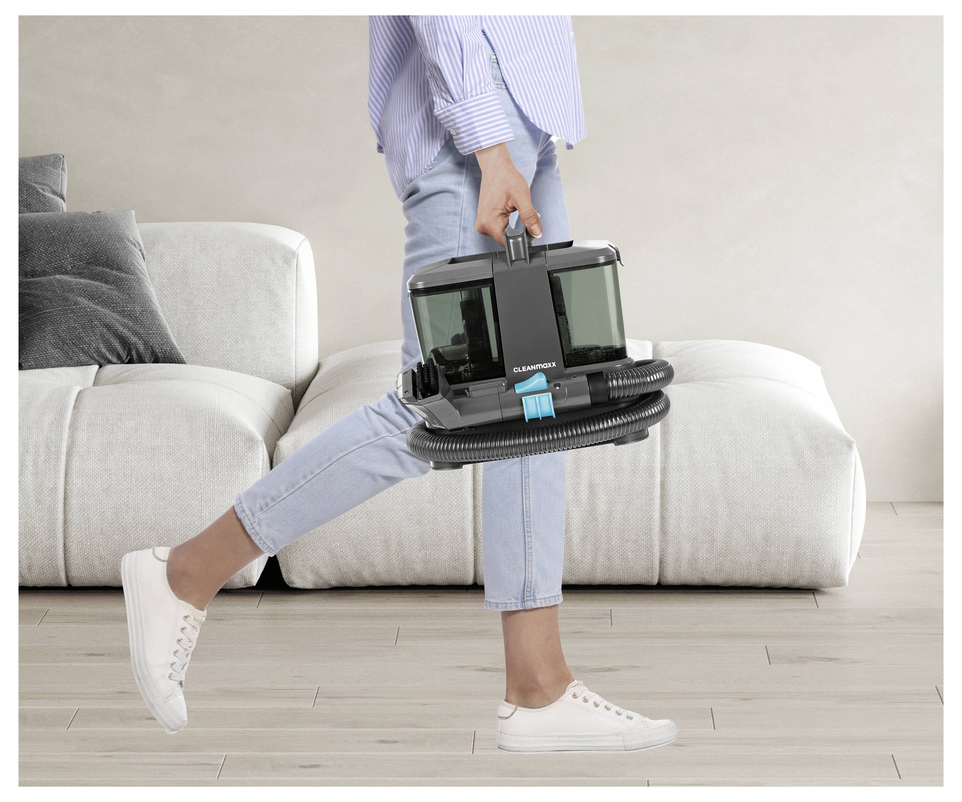 Een persoon in casual kleding loopt binnenshuis en draagt een compact, modern stofzuigerapparaat naast een witte bank in een woonkameromgeving.