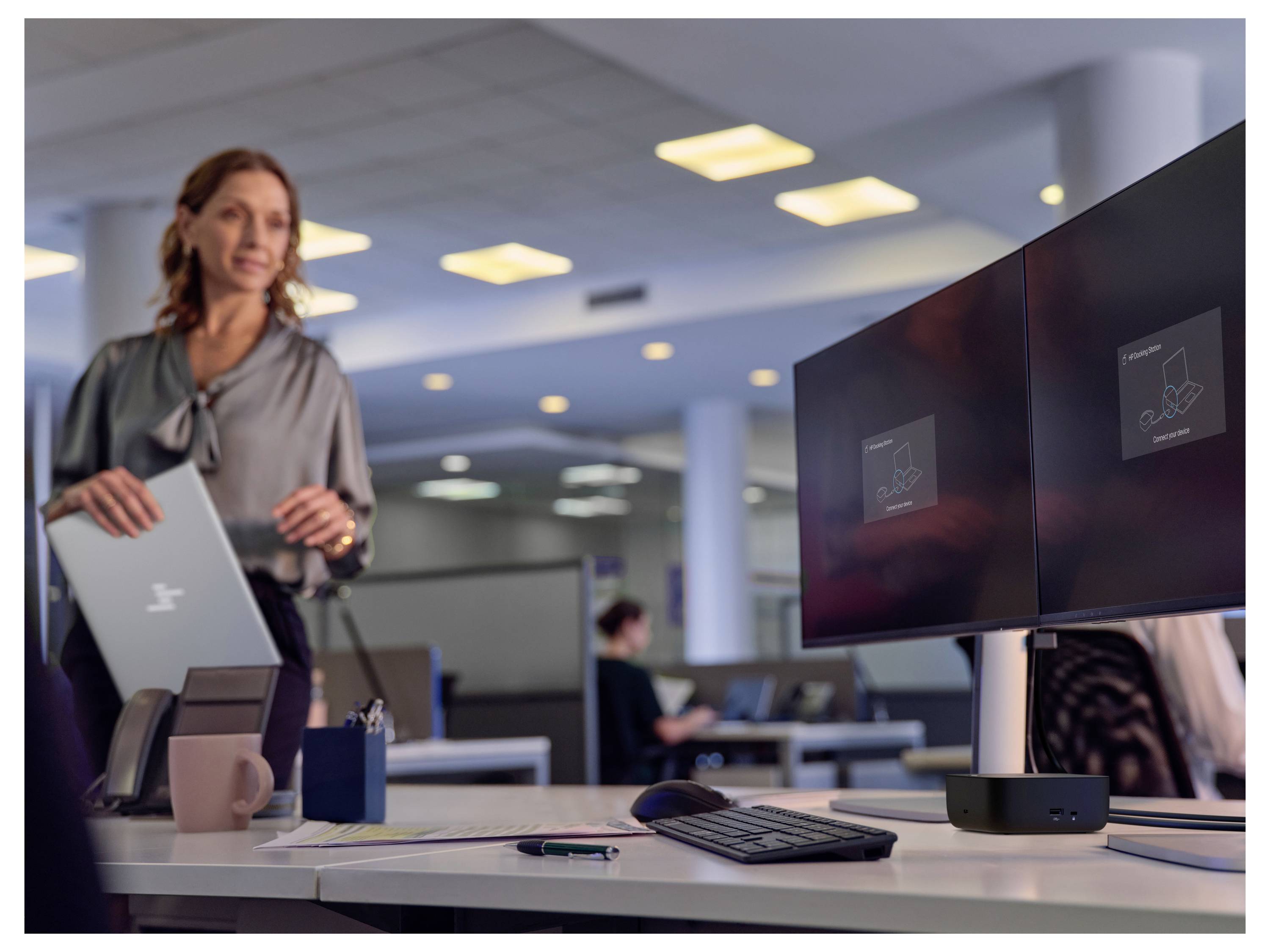 Een vrouw houdt een laptop vast in een modern kantoor. Twee schermen op een bureau tonen digitale inhoud. Kantoorsverlichting en andere werknemers op de achtergrond.