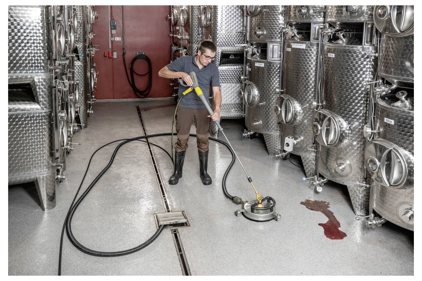 Een persoon die laarzen draagt, gebruikt een vloerreiniging machine in een industriële omgeving met metalen tanks. Er is een rode vloeistofmorser in de buurt.