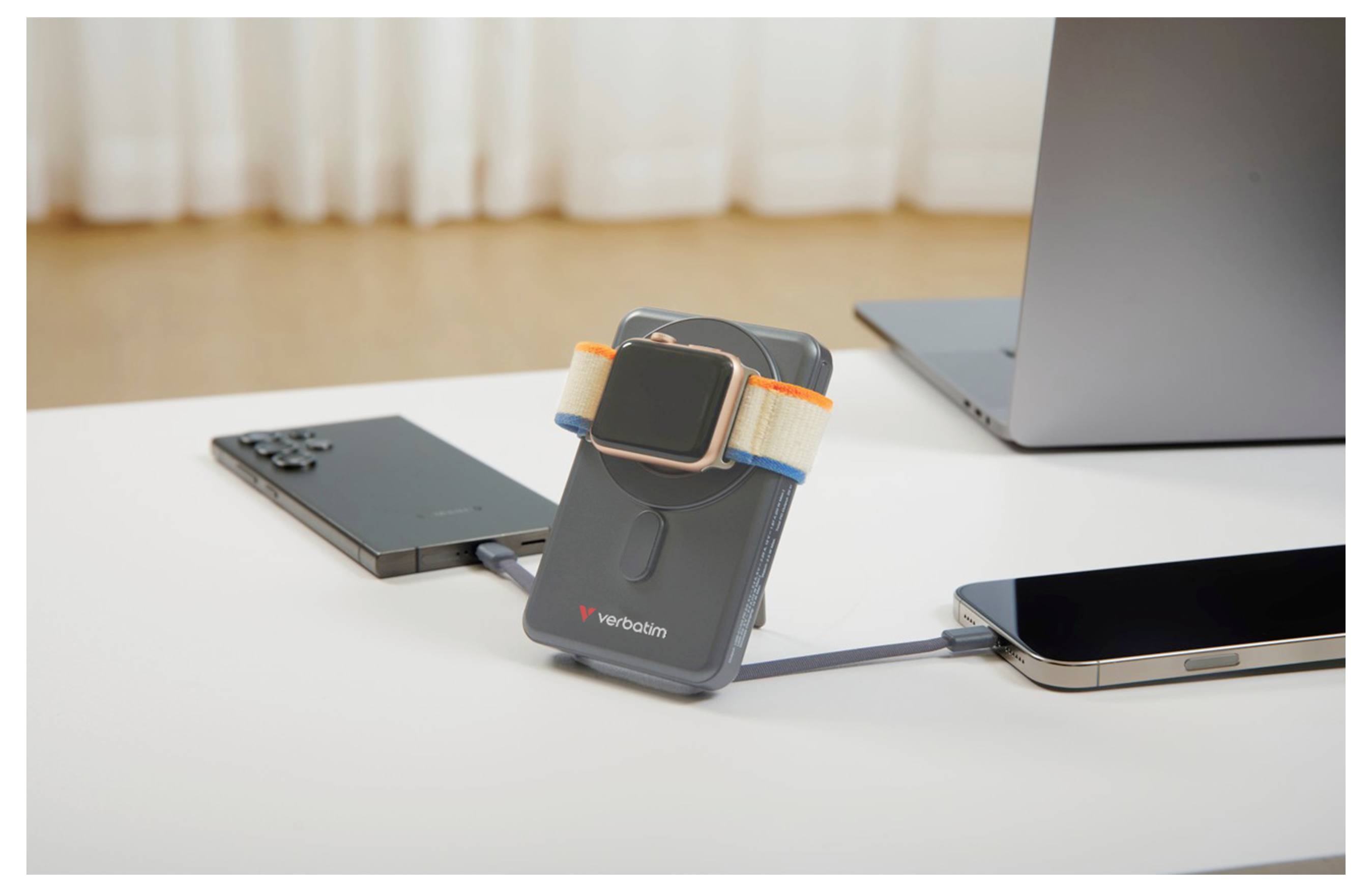 Een smartwatch die oplaadt op een draagbare powerbank, verbonden met twee smartphones op een wit bureau, met een laptop op de achtergrond.