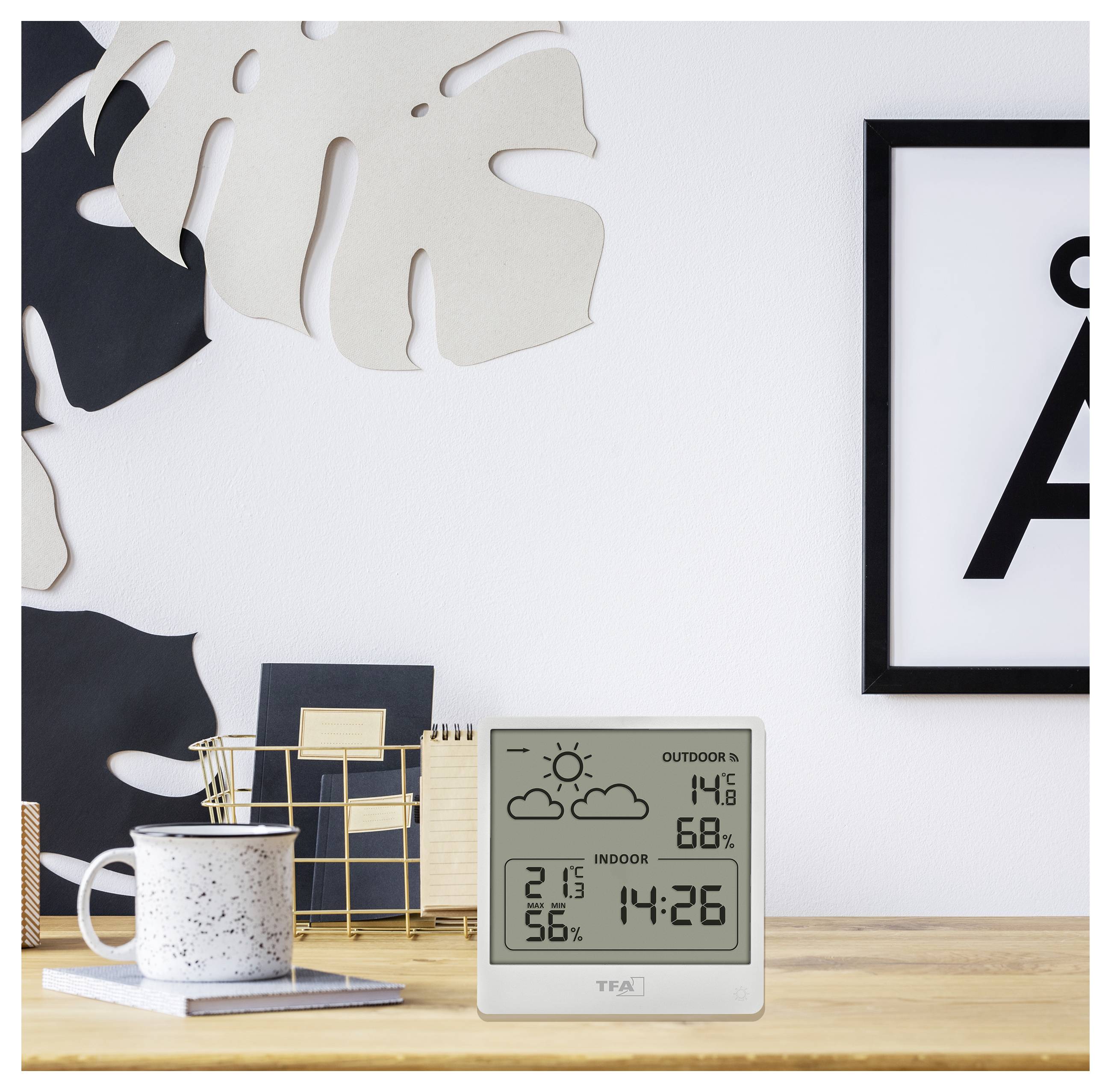 Een digitale binnen-buitenthermometer geeft weer: Buiten 14,8°C, 68% luchtvochtigheid; Binnen 21,3°C, 56% luchtvochtigheid, tijd 14:26, geplaatst op een houten tafel nabij decoratie.