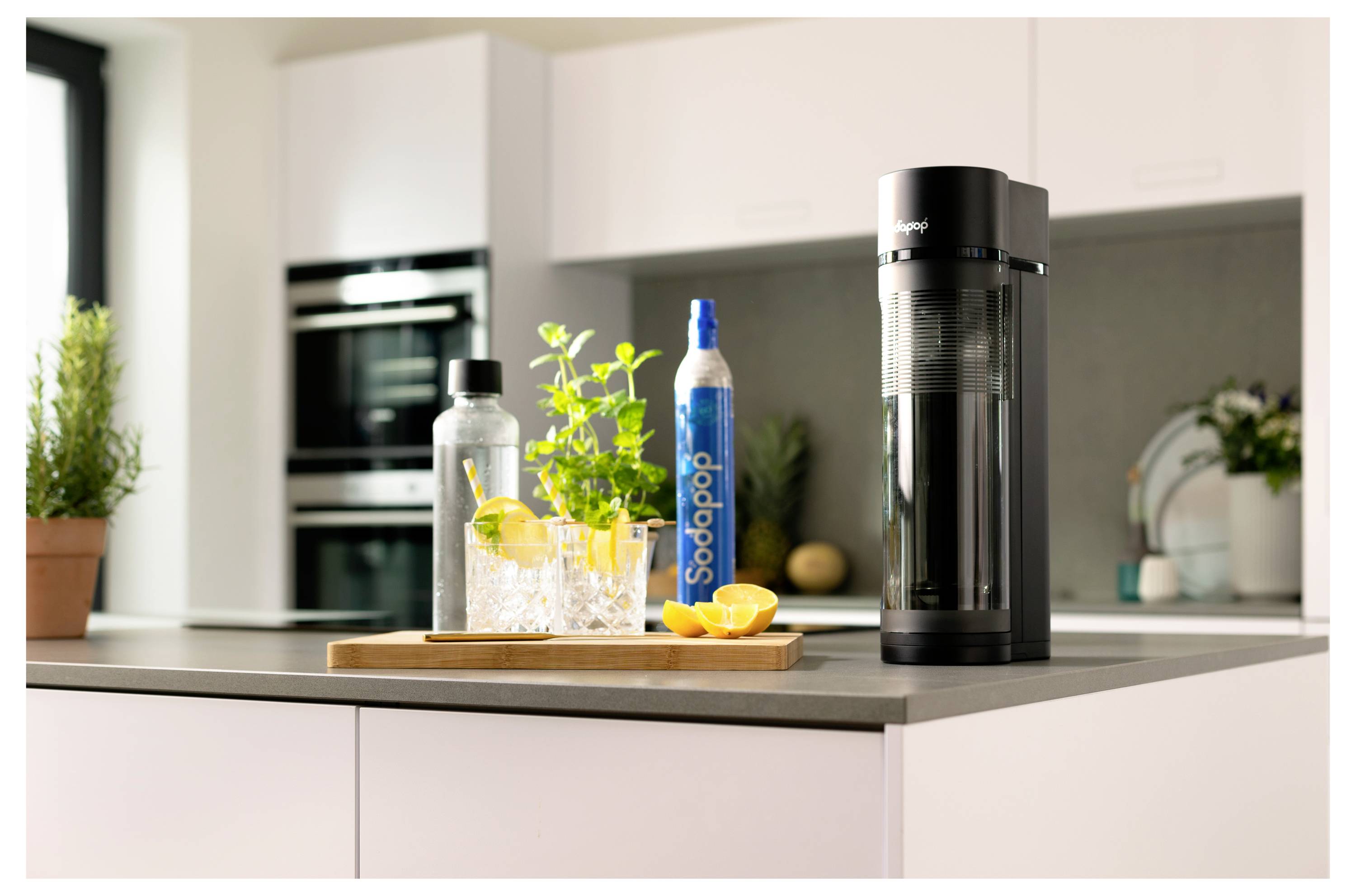 Een aanrecht in de keuken met een sodamaker, een fles met het label 'Sodapop,' glazen met citroen en kruiden, en een moderne keuken op de achtergrond.