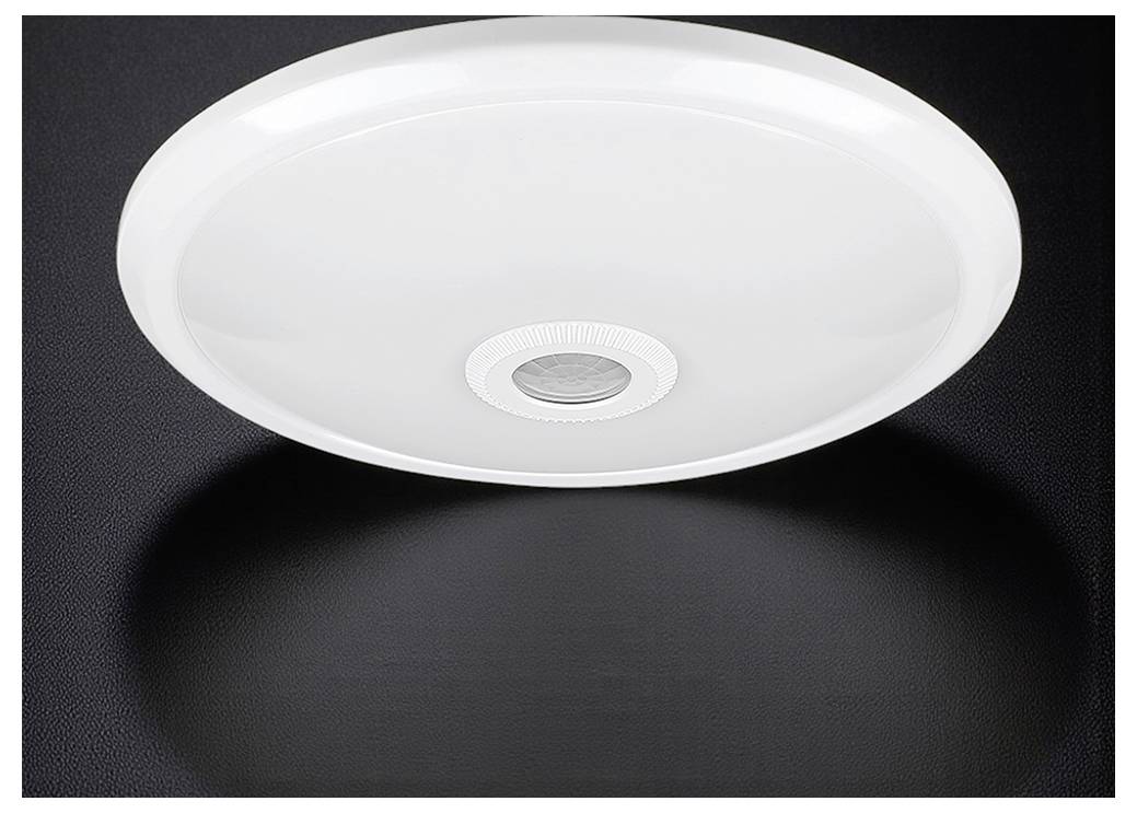 Een rond, wit plafondlamp met een centraal sensor, gemonteerd op een donker oppervlak, dat een zachte schaduw eronder werpt.