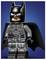Een LEGO-figuur van een superheld met een zwart pak, vleermuis-embleem op de borst en cape, staand tegen een blauwe achtergrond.