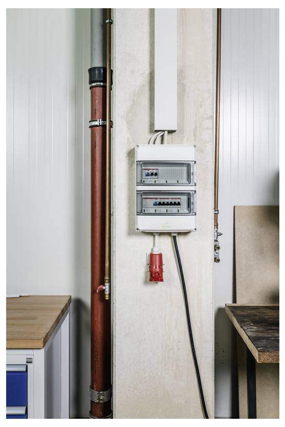 Elektrisch paneel en rode pijp geïnstalleerd op een onafgewerkte binnenmuur tussen werkbanken, met een kabel die omlaag loopt naar een rood contactdoosje.