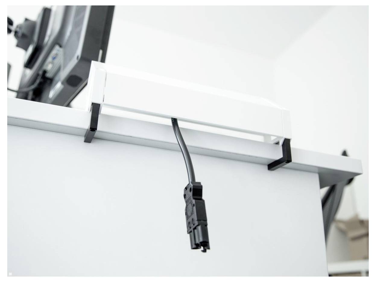 Een zwarte kabel die met clips is bevestigd, hangt aan de onderkant van een wit bureau, naast een monitor op een gekantelde arm in een modern kantooromgeving.