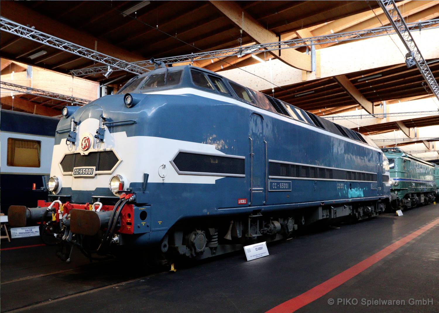 Een blauwe vintage locomotief tentoongesteld in een binnenruimte op sporen, gelabeld '0165900'. De omgeving suggereert een spoorwegmuseumomgeving.