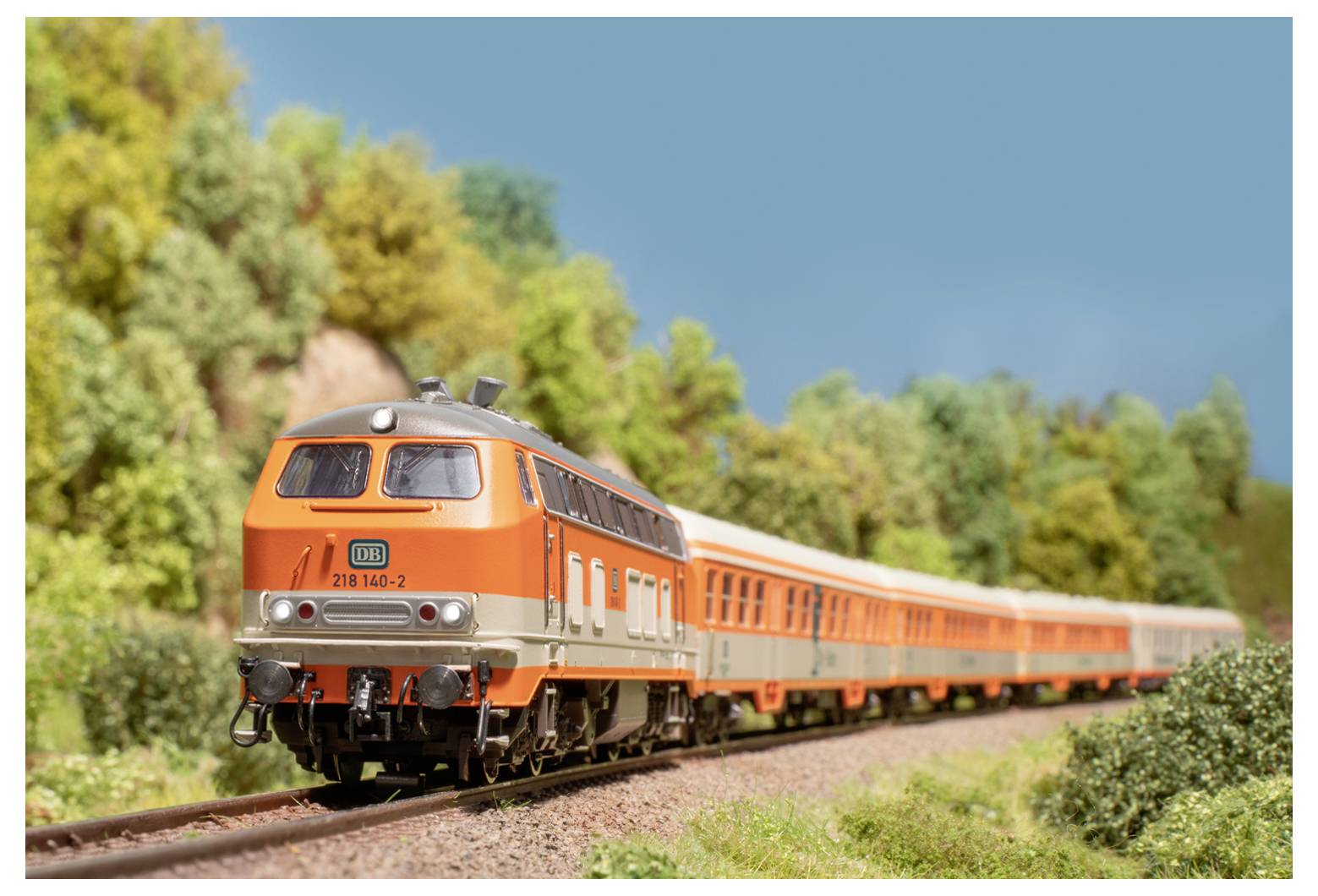 Een oranje trein reist op een spoorweg door een schilderachtig landschap met bomen en een heldere hemel.
