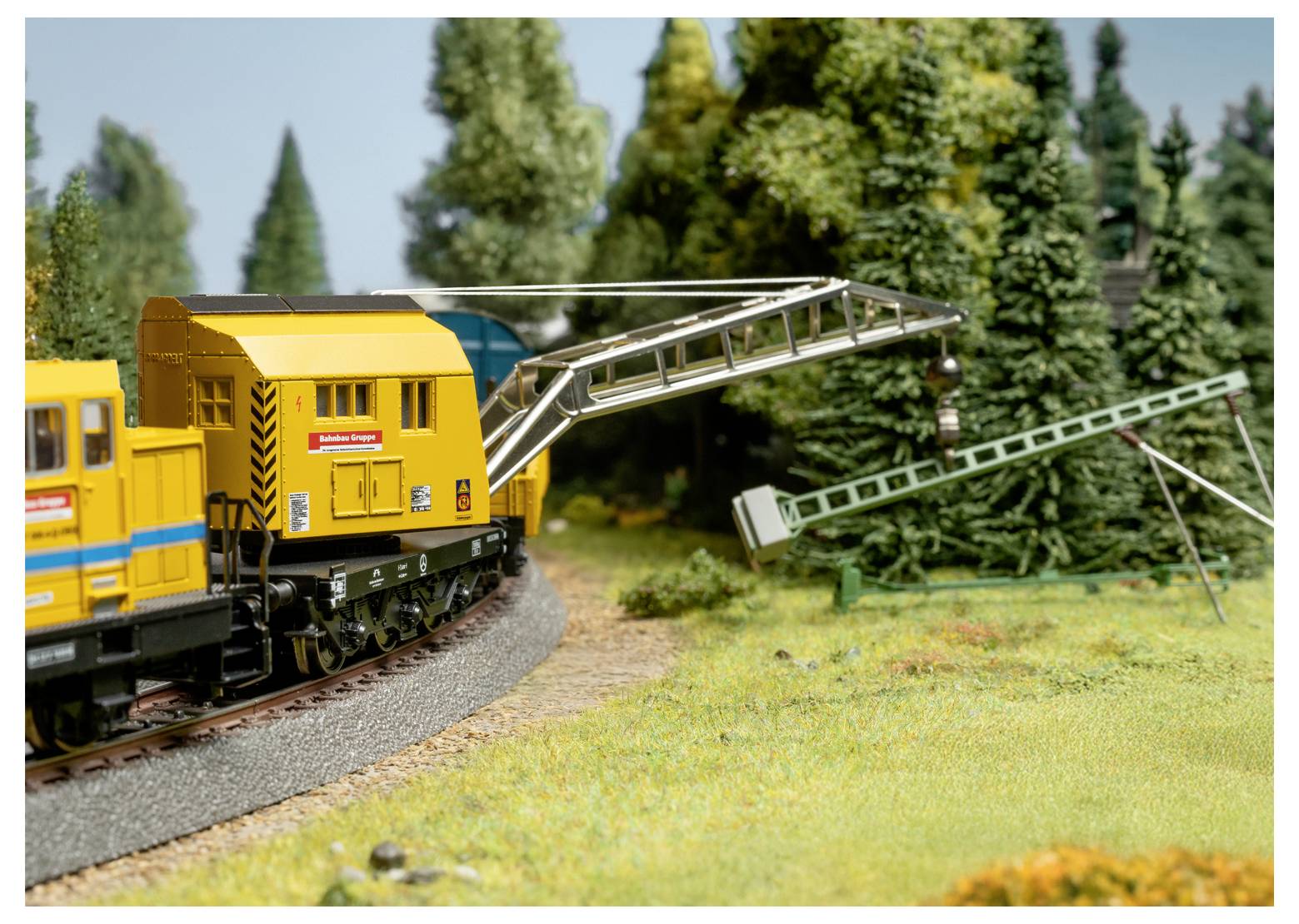 Een modelspoorbaan-scène met een onderhoudstrein met gele wagons die over een gebogen spoor rijdt naast een bosrijk gebied en kraanuitrusting.
