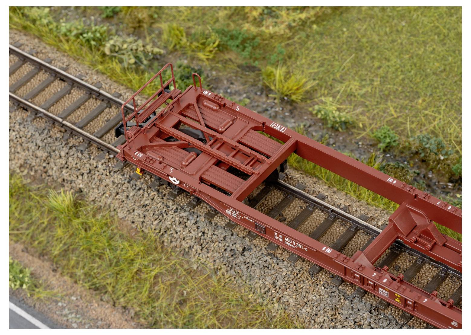 Een modeltreinplatformwagon met realistische details staat op een spoor omgeven door gras en grind, met een scène uit een miniatuurrailwayopstelling.