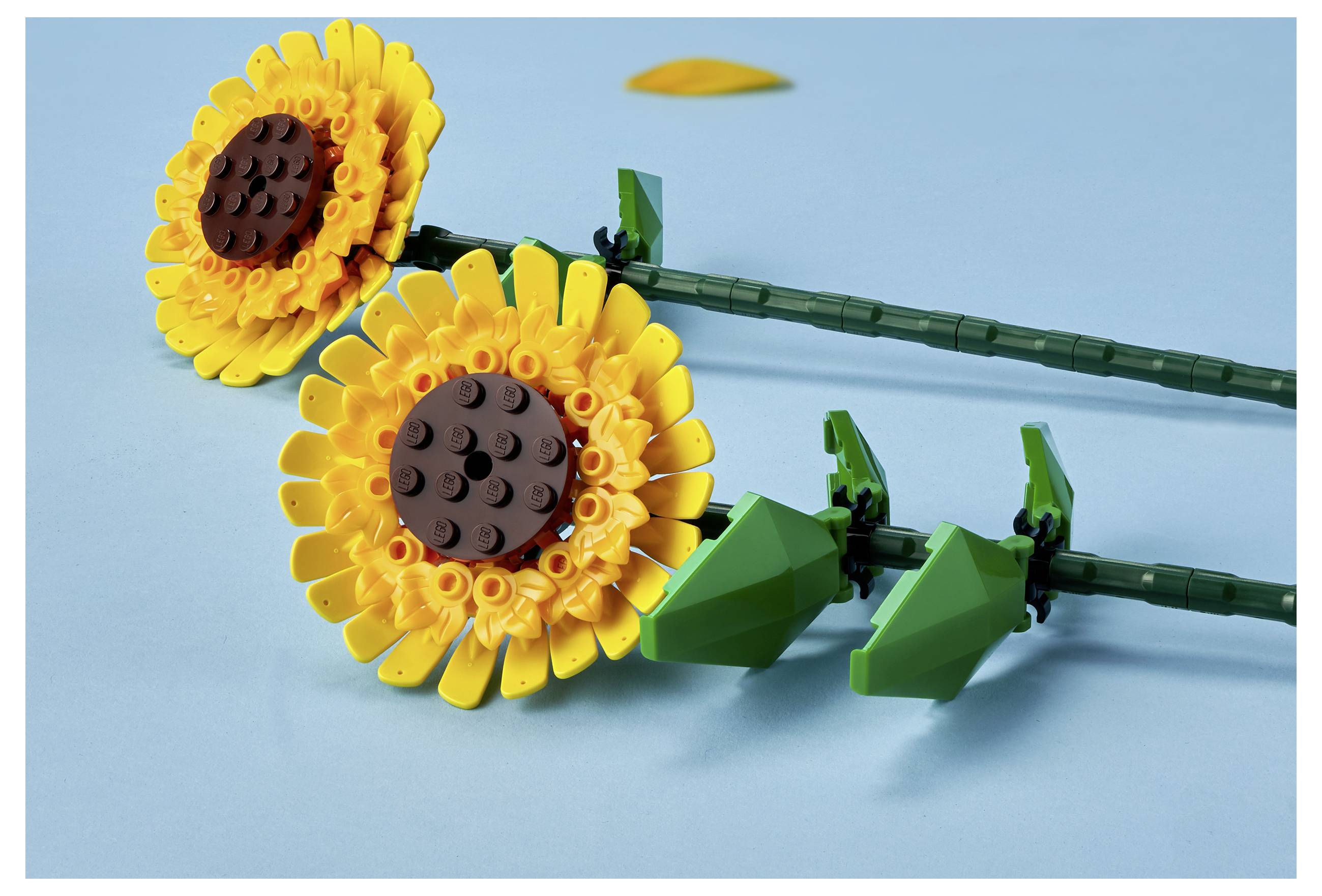 Twee LEGO zonnebloemen met gele bloemblaadjes en bruine centra liggen op een lichtblauwe achtergrond en tonen gedetailleerde ontwerpen van stengels en bladeren.