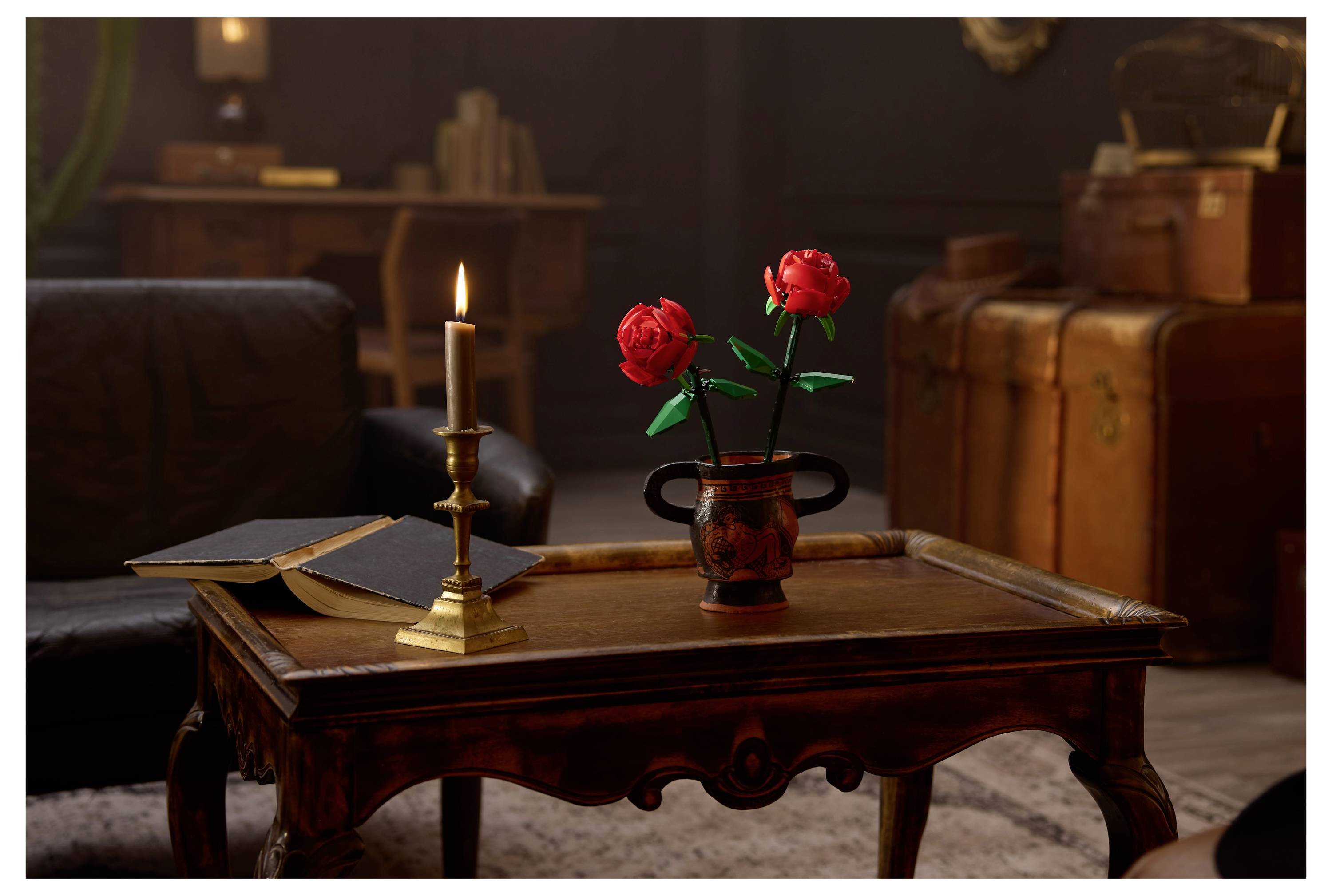 Een zwak verlicht vertrek met een houten tafel, voorzien van een brandende kaars, een opengeslagen boek en een vaas met rode rozen. Vintage koffers op de achtergrond.